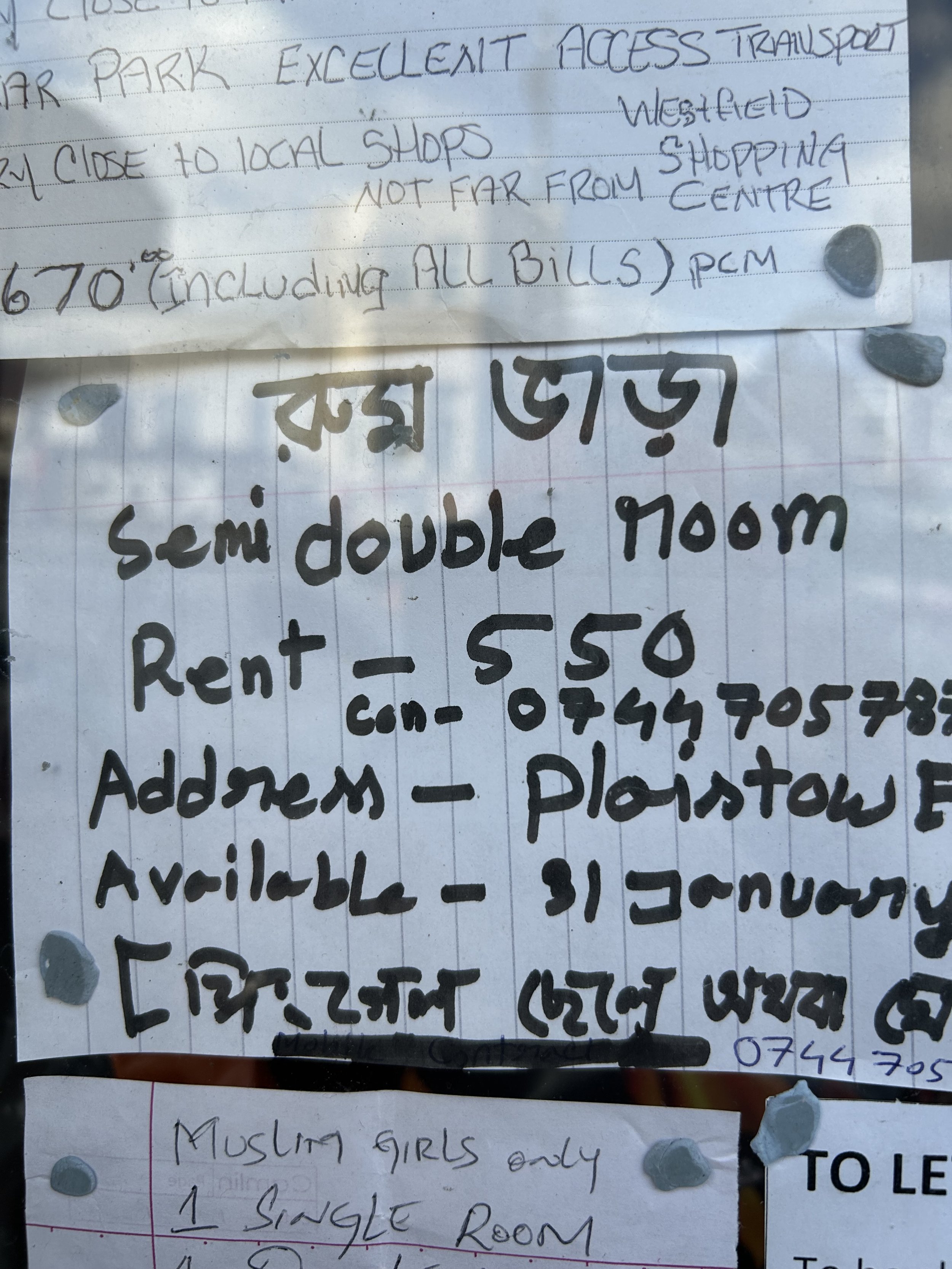 A handwritten sign in English and another language, advertising a semi-double room for rent in a building called 'Max Guest'. The sign details rent cost, contact number, address, and availability. There are small stones attached to the paper.