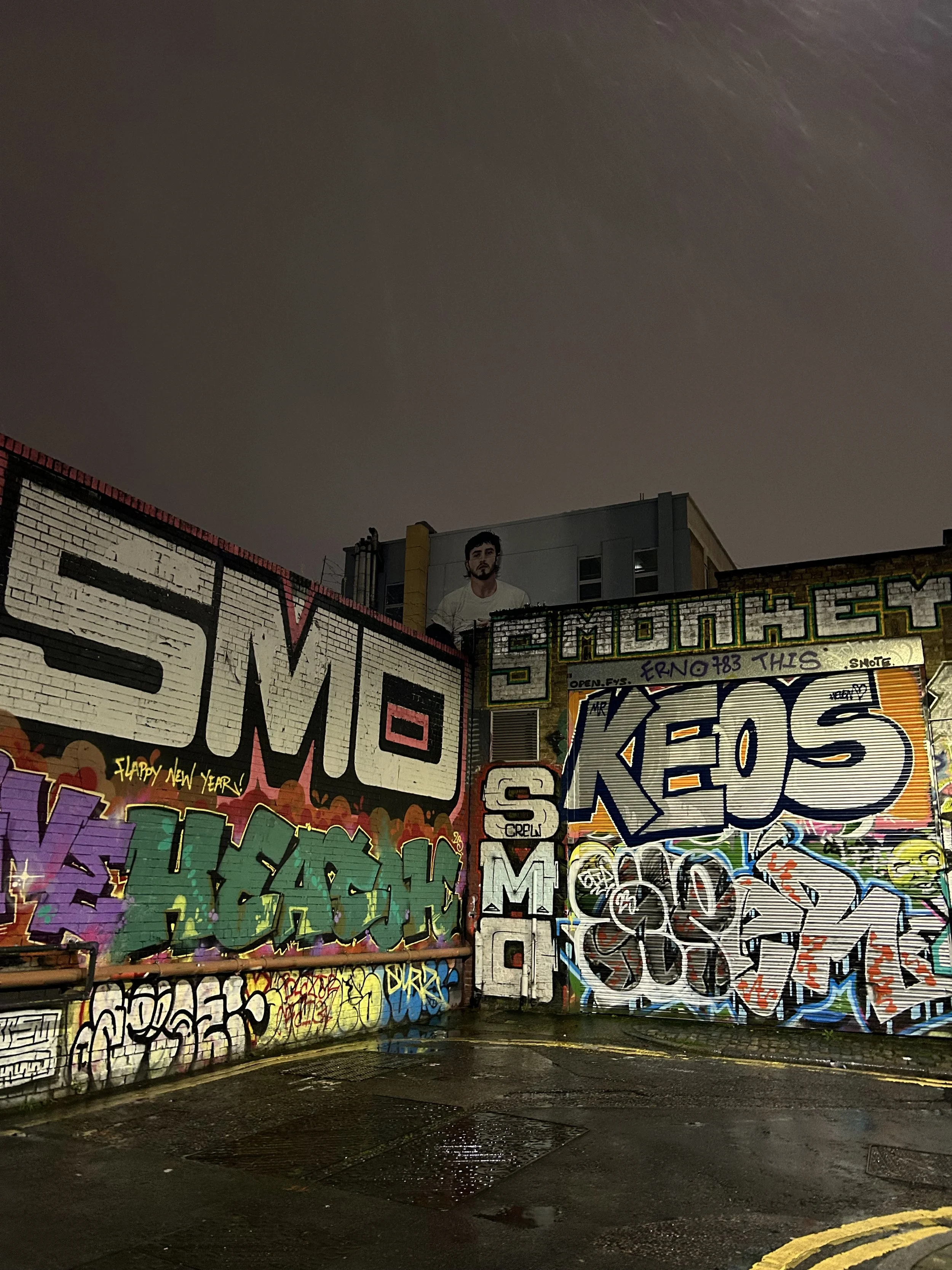 Nighttime view of graffiti-covered walls with large colorful tags and murals, including the words 'SMOKE' and 'KEOS,' and a person’s portrait mural can be seen high on the building in the background.