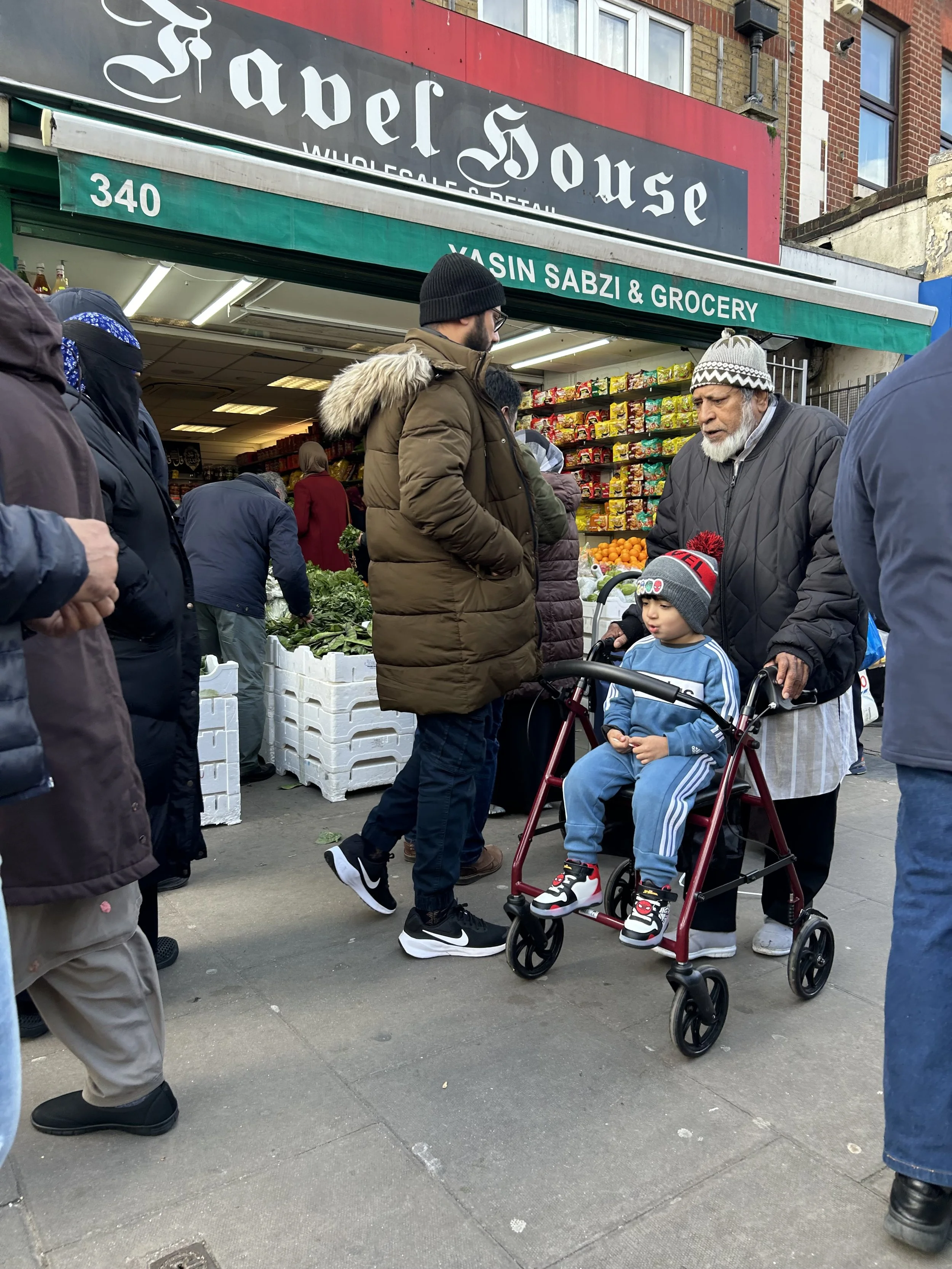 A busy outdoor street market with people shopping for groceries. A man in a brown coat and black beanie stands next to an elderly man with a white beard, who is pushing a young boy in a red walker. The store sign reads "Yasin Sabzi & Grocery" with pr