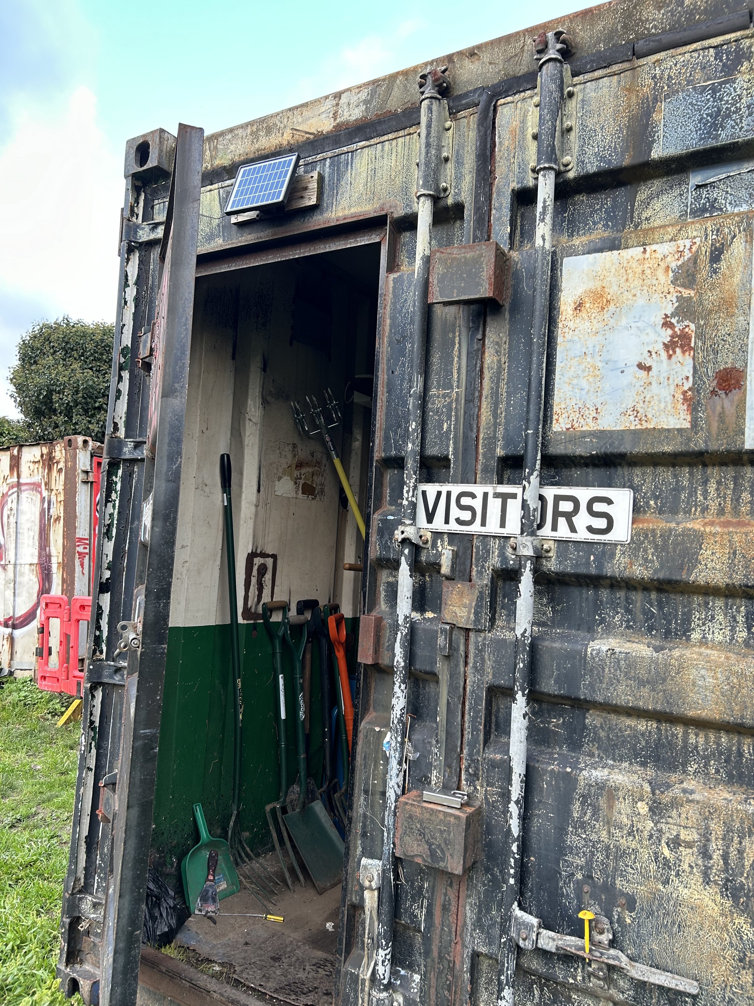 rusty metal storage container with open door revealing gardening tools inside, a solar panel mounted on the exterior, and a 'VISITORS' sign on the door.