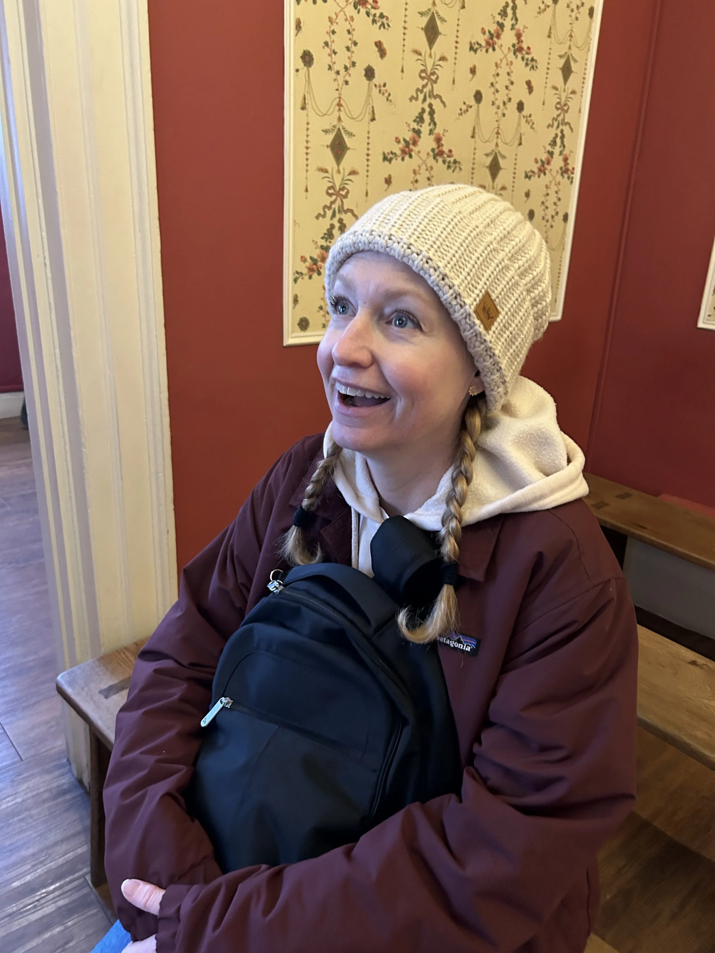 A woman wearing a beige knit beanie, maroon jacket, and a backpack, sitting indoors with a surprised and happy expression, against a wall with vintage floral wallpaper.