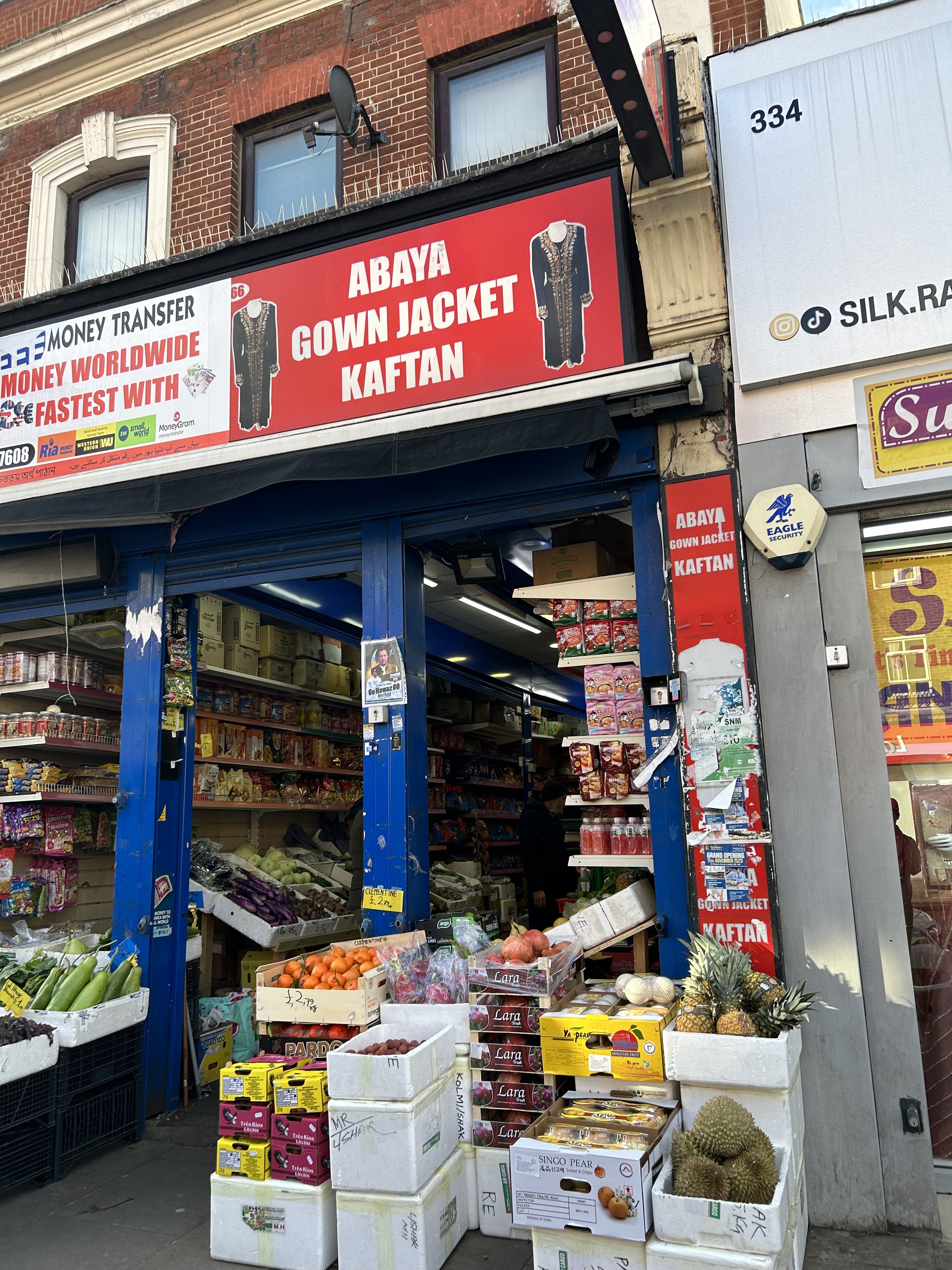 View of a small grocery store with a sign advertising Abaya gown jacket and Kaftan. The store has produce including cucumbers, eggplants, oranges, pomegranates, pineapples, and durians displayed outside. Shelves inside are stocked with various packag
