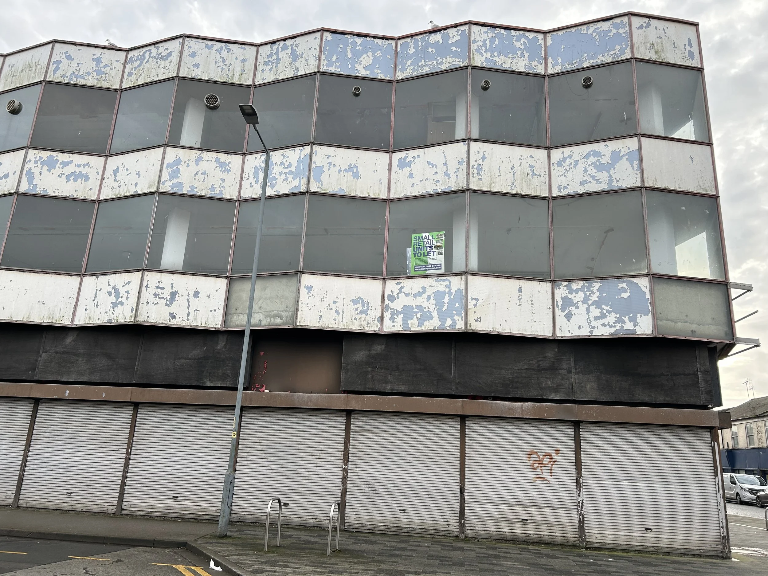 An abandoned multi-story building with broken, peeling blue and white paint on the exterior walls, large empty windows, a closed shutter on the ground floor, graffiti, a streetlamp, and a cloudy sky.