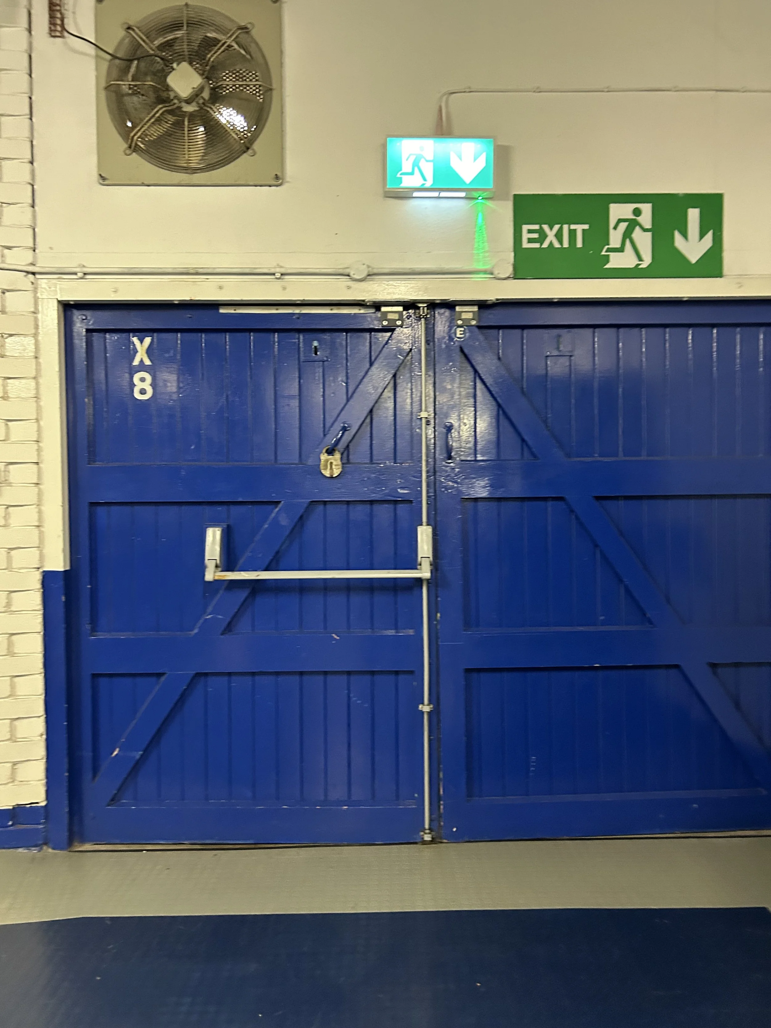 Blue barn-style door with a vertical lock, located under exit signs with green and blue emergency exit indicators above.