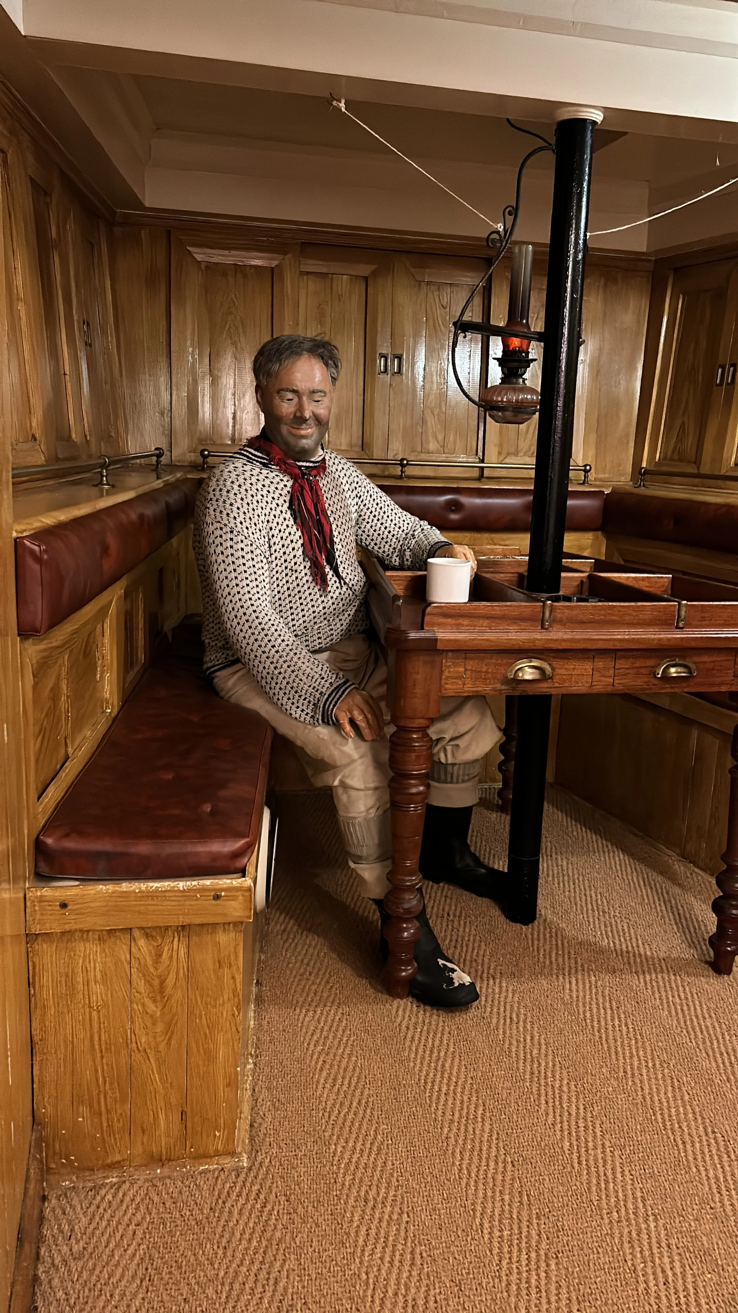 A life-sized figure of a man dressed as a cowboy, sitting in a wooden booth at a themed restaurant, with a cup on the table and a vintage oil lantern hanging above.