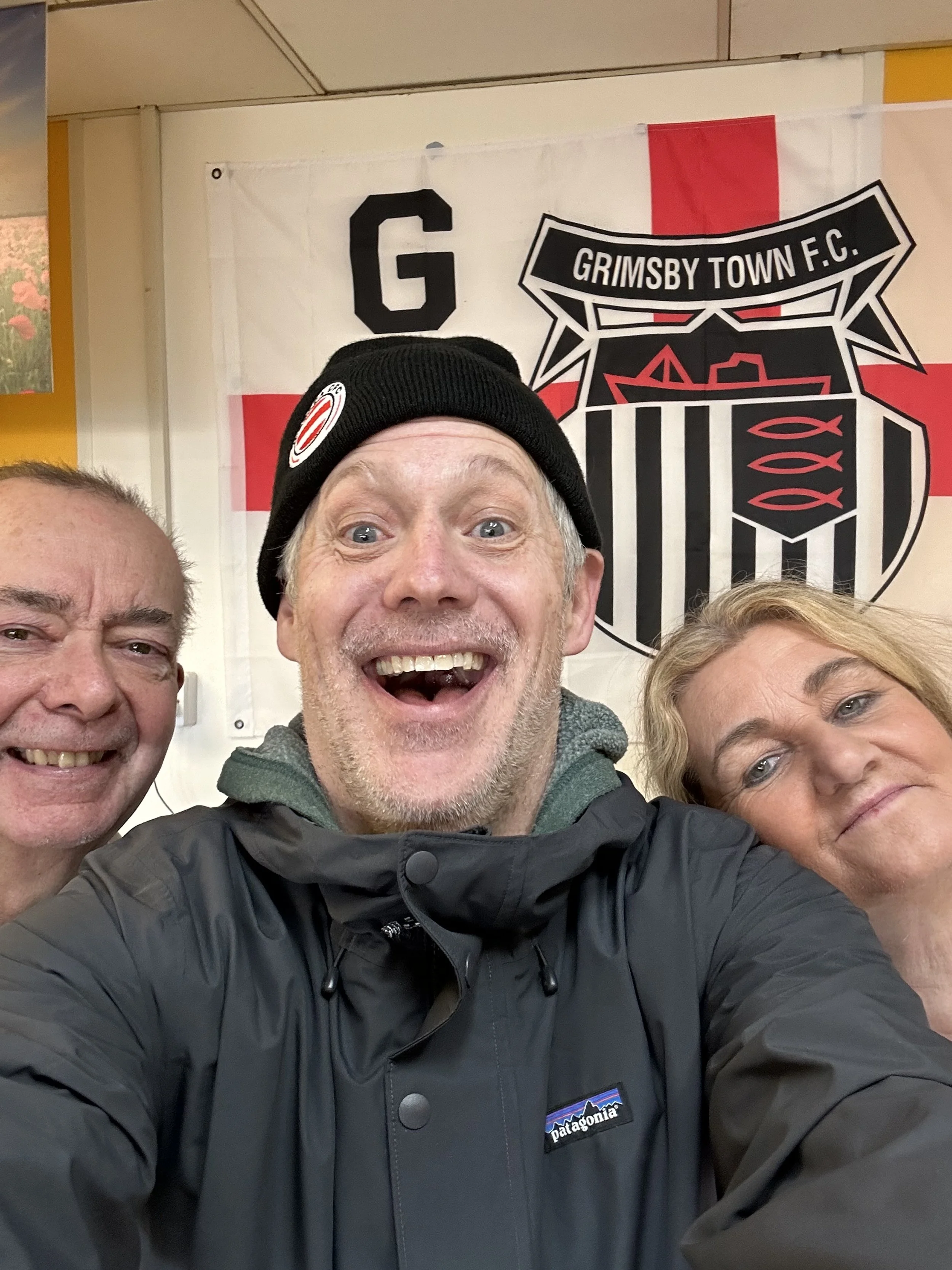 Group of three people taking a selfie in front of a Grimsby Town F.C. flag, smiling happily.