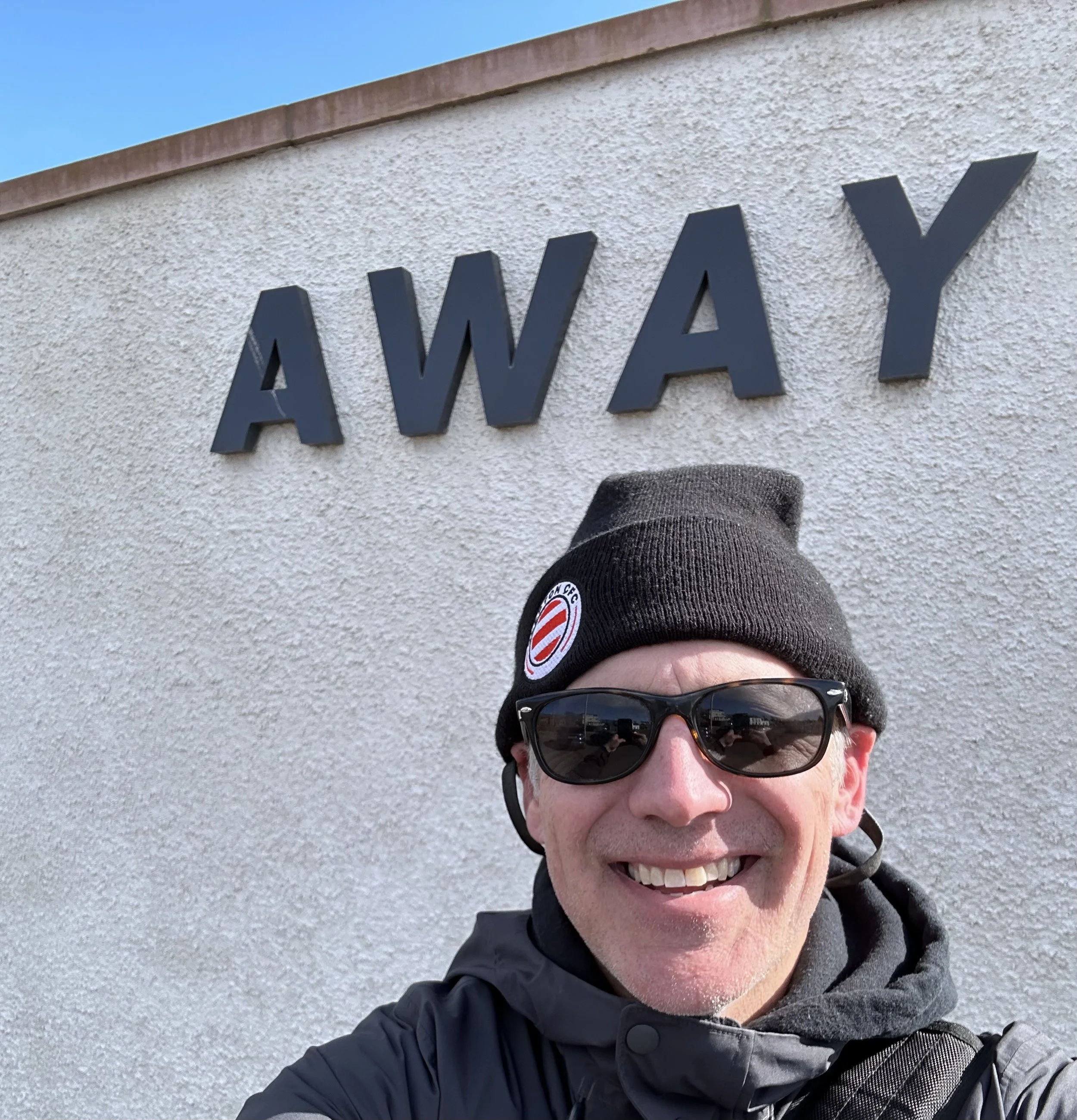 Smiling man wearing sunglasses, black beanie with a logo, and jacket taking a selfie in front of a wall with the word 'AWAY' in large black letters.