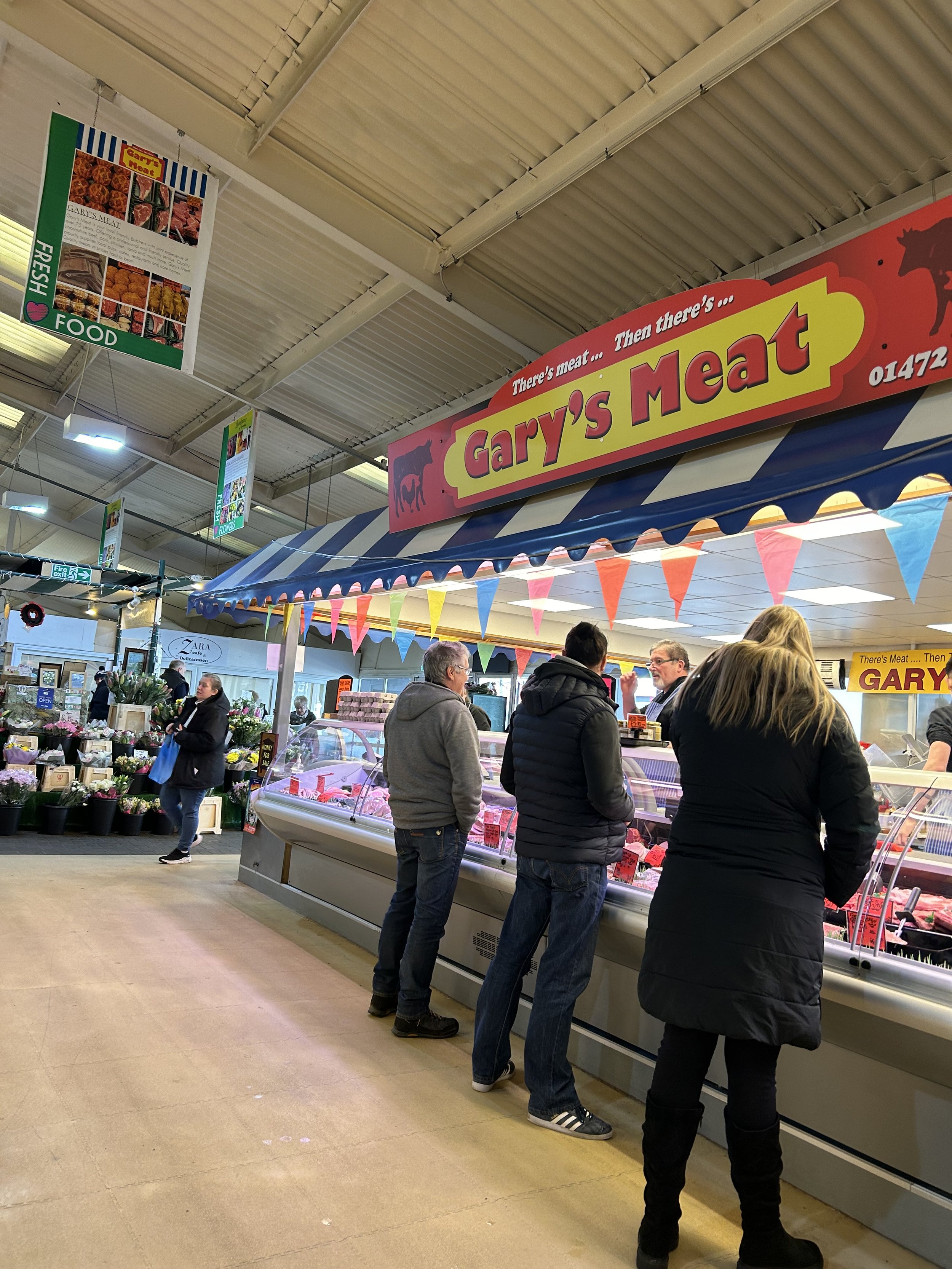 People shopping for meat at Gary's Meat inside a market, with a sign saying 'There’s meat... Then there’s Gary’s Meat'.