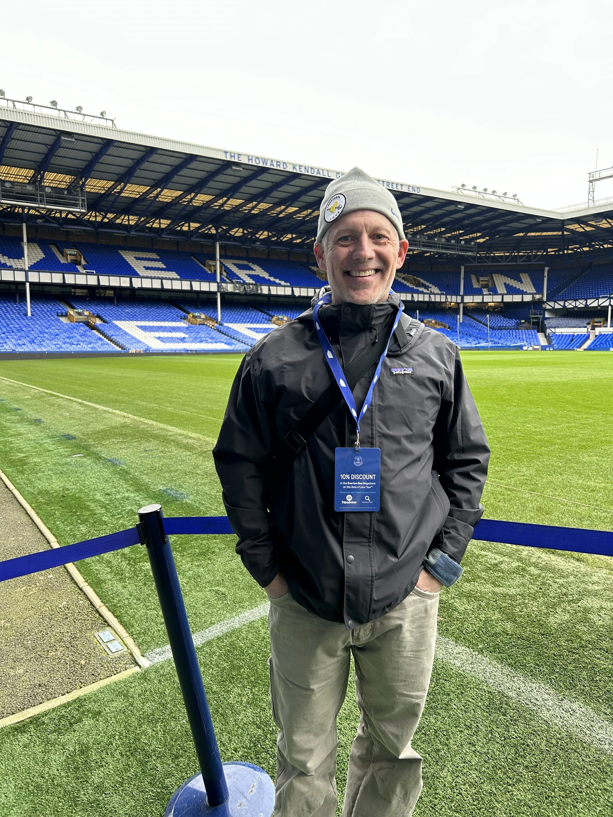 A smiling man wearing a gray beanie and black jacket standing on a football field at a stadium. He has a lanyard with a blue discount card around his neck. The stadium has blue seats and a sign that reads 'THE HOWARD KENDALL' at the top.