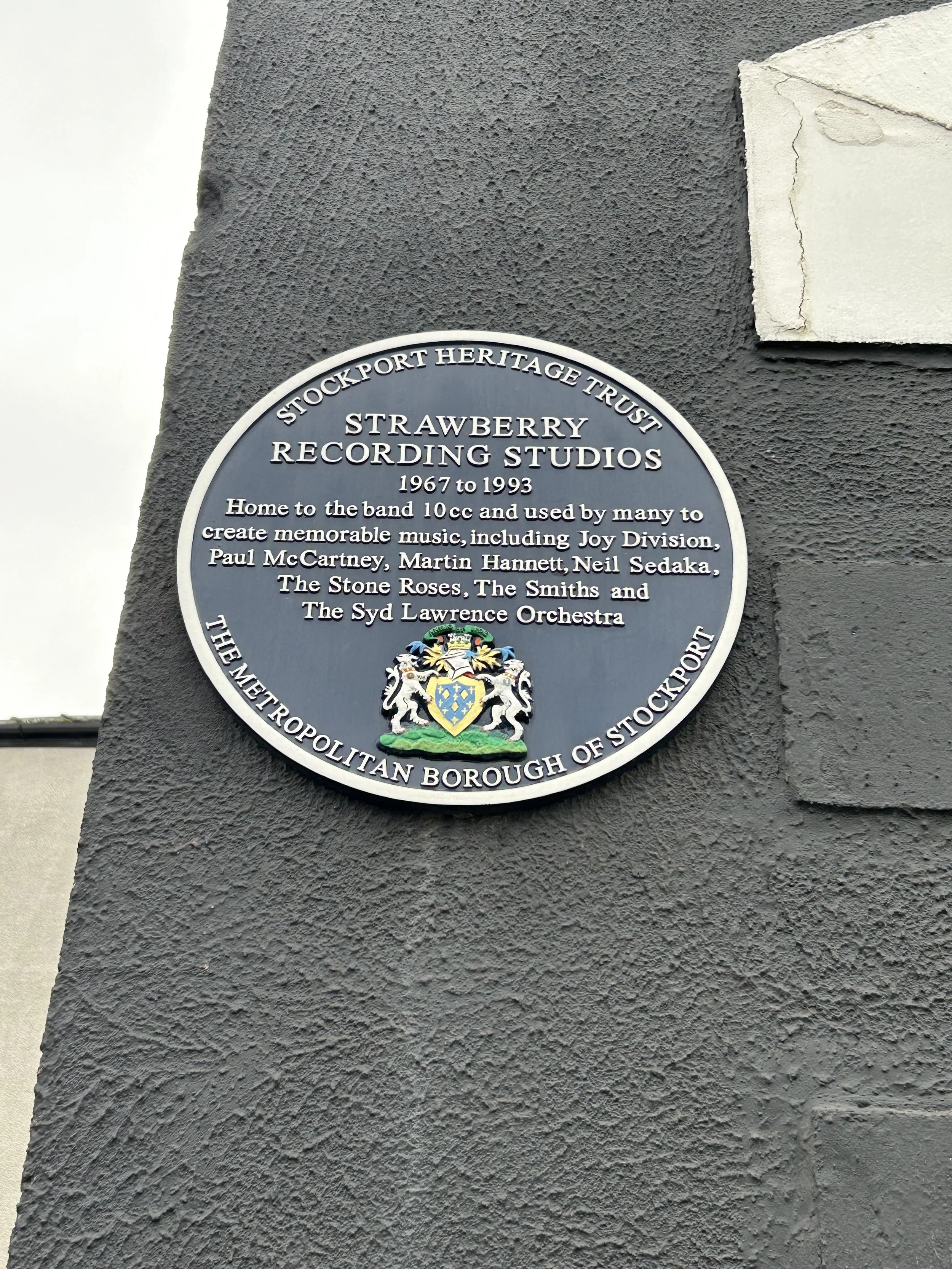 Black oval plaque on a gray wall commemorating Strawberry Recording Studios, home to famous bands from 1967 to 1993, with a colorful coat of arms at the bottom.