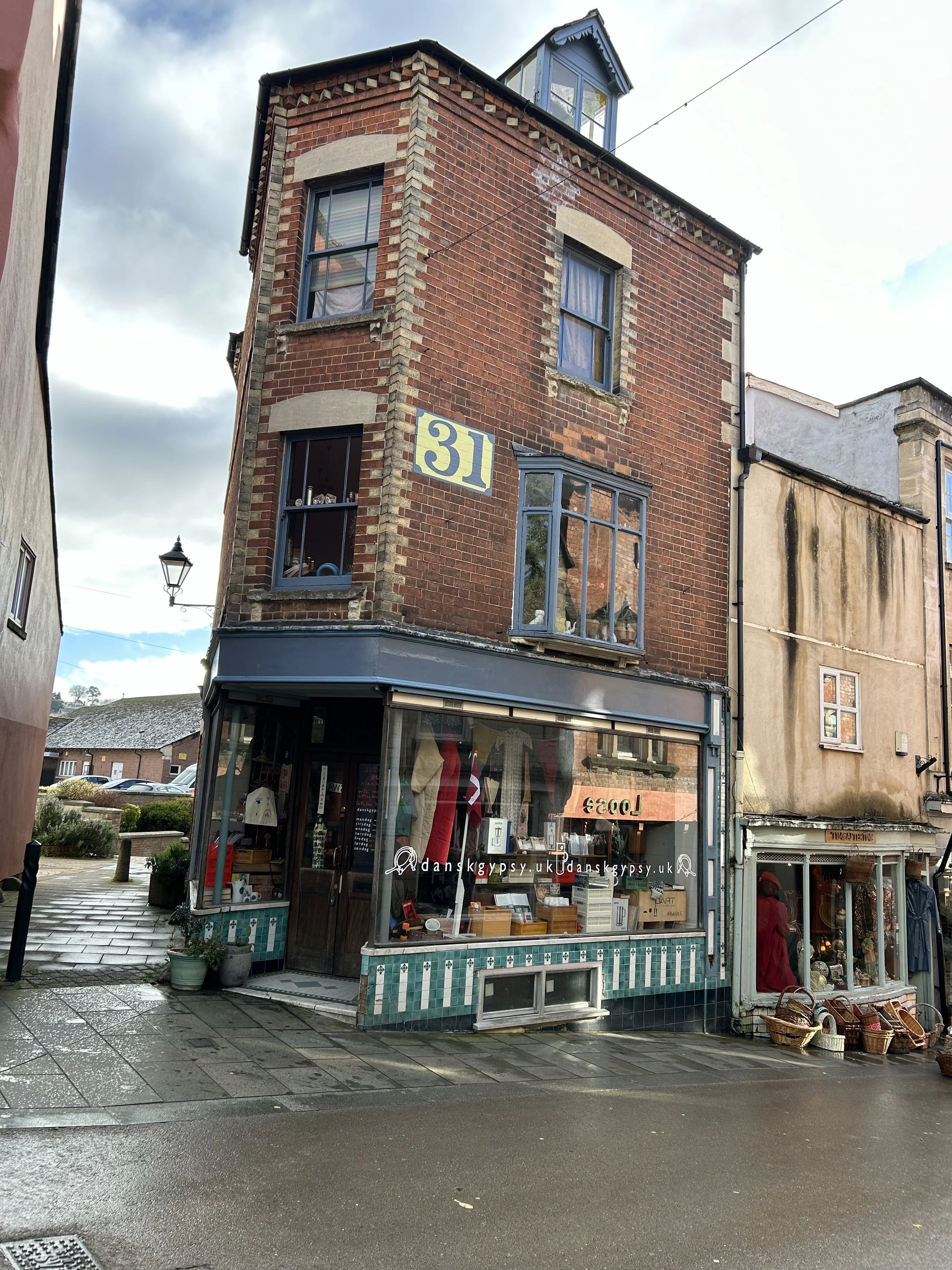 A three-story brick building with a storefront on the ground floor and the number 31 on the wall. The storefront has large glass windows displaying clothing and accessories, with decorative green and white tiles at the bottom. Potted plants are outsi