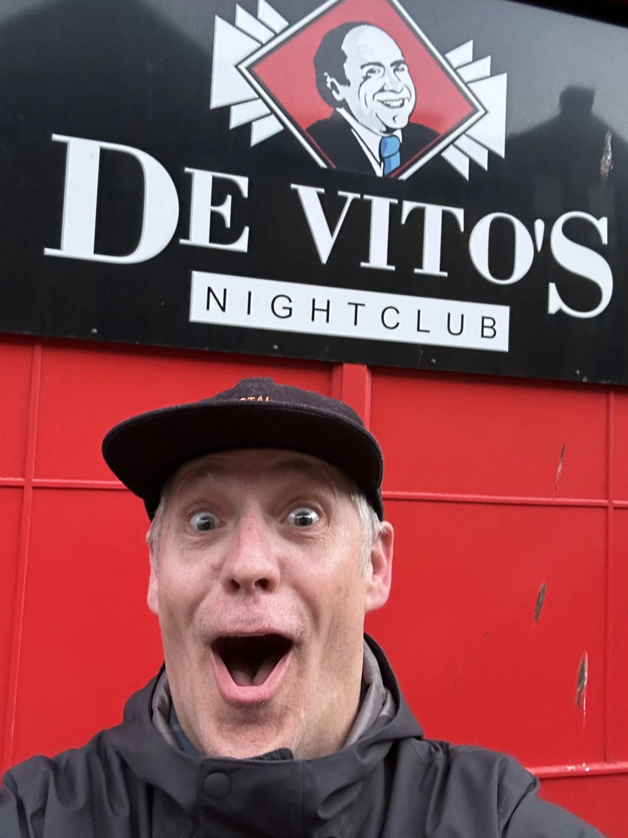 A man taking a selfie in front of a black and red sign for De Vito's Nightclub. The man is wearing a black cap and a black jacket, with an excited expression and open mouth.