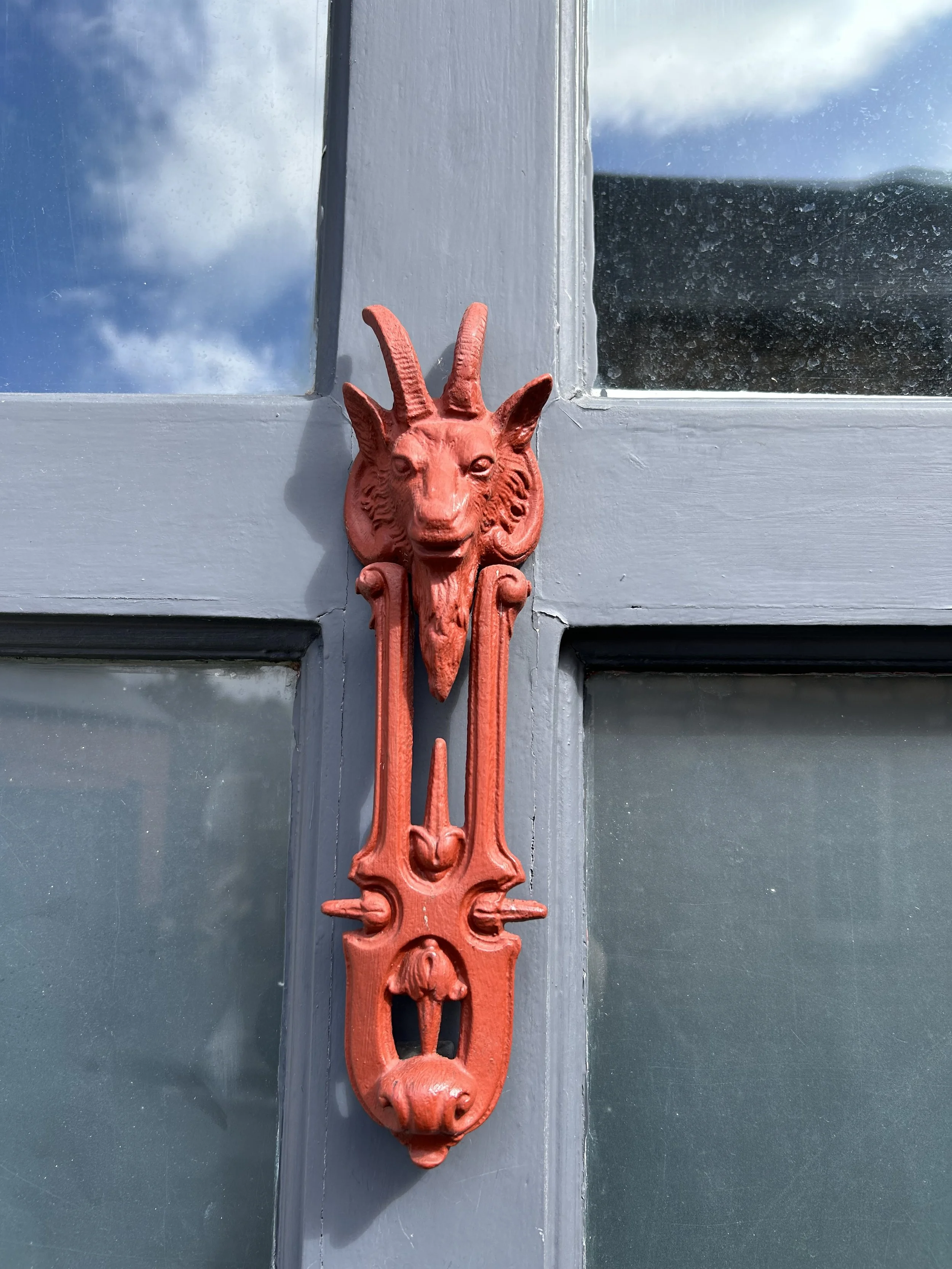 Close-up of a ornate, red door knocker with a goat's head design mounted on a gray door with glass panes reflecting clouds and sky.