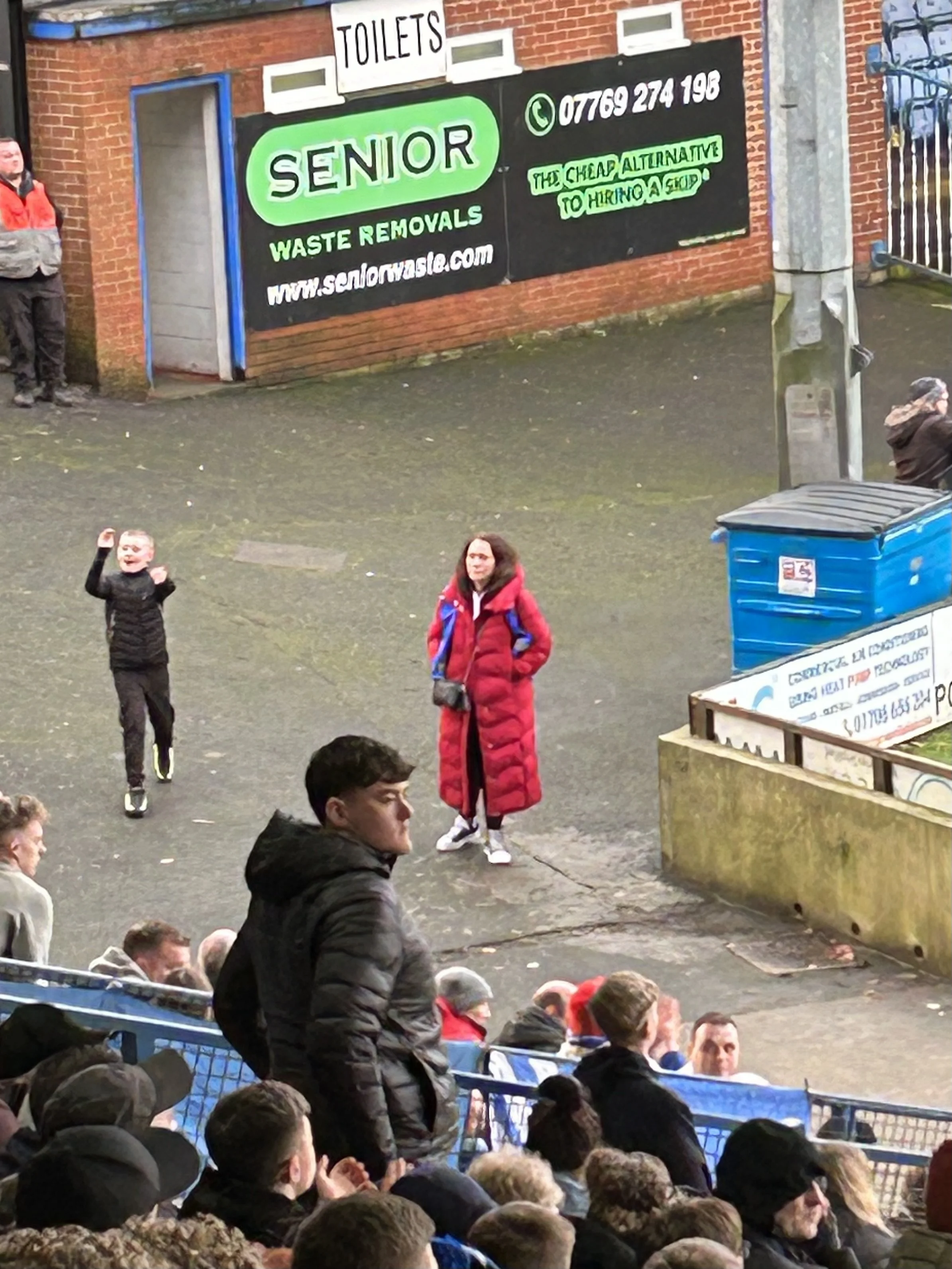 People sitting and standing outside viewing an event, with a woman in a red coat and a young boy wearing a black jacket in the foreground; a large billboard advertising senior waste removal services is visible on the brick wall behind them.