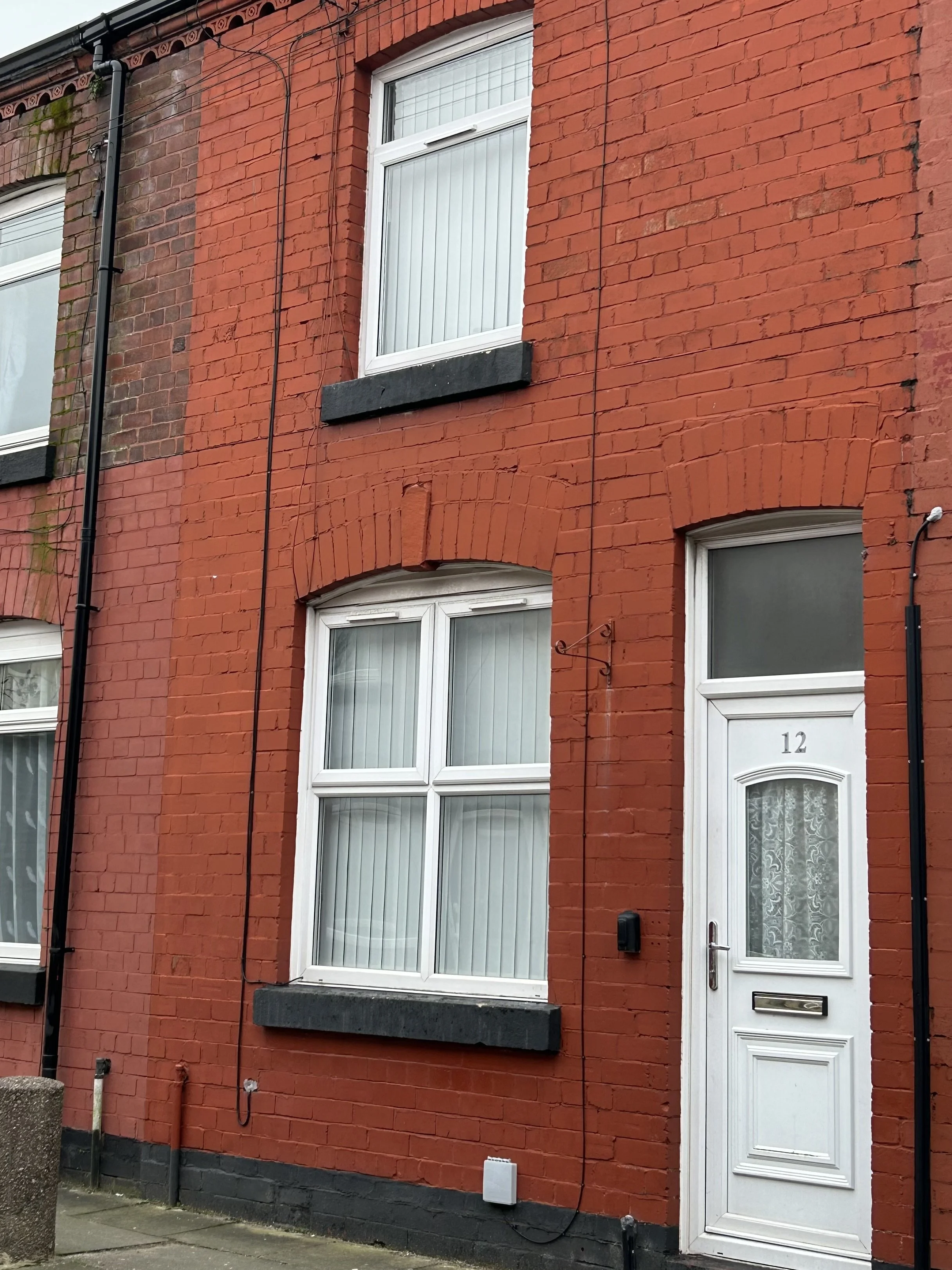 Red brick house with white window frames and door number 12, curtain in the ground floor window, black trim on windows, black electrical wires, and drain pipes.