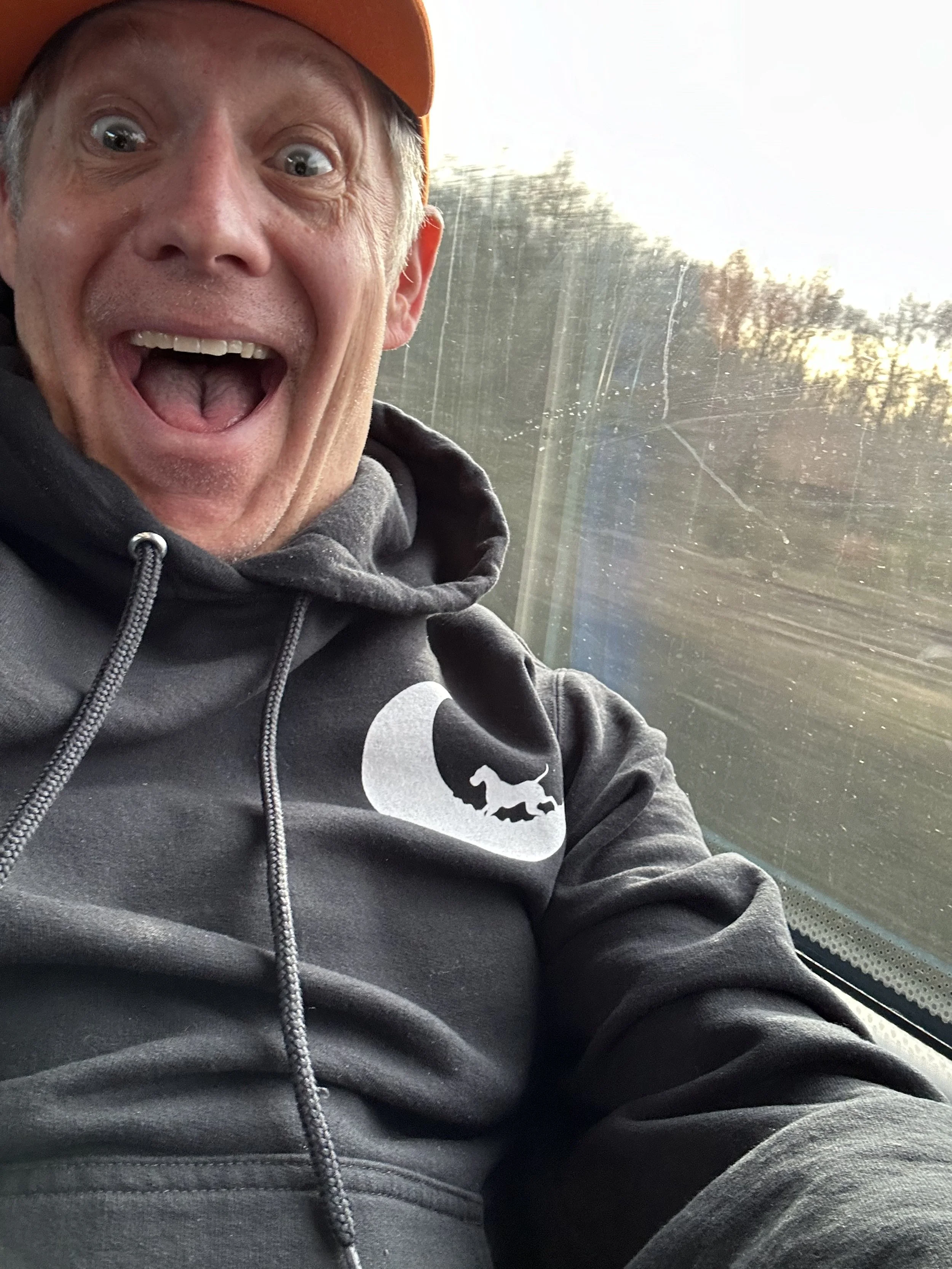 A man wearing a gray hoodie with a logo of a dog in front of a window, smiling widely with an open mouth, and wearing an orange cap.