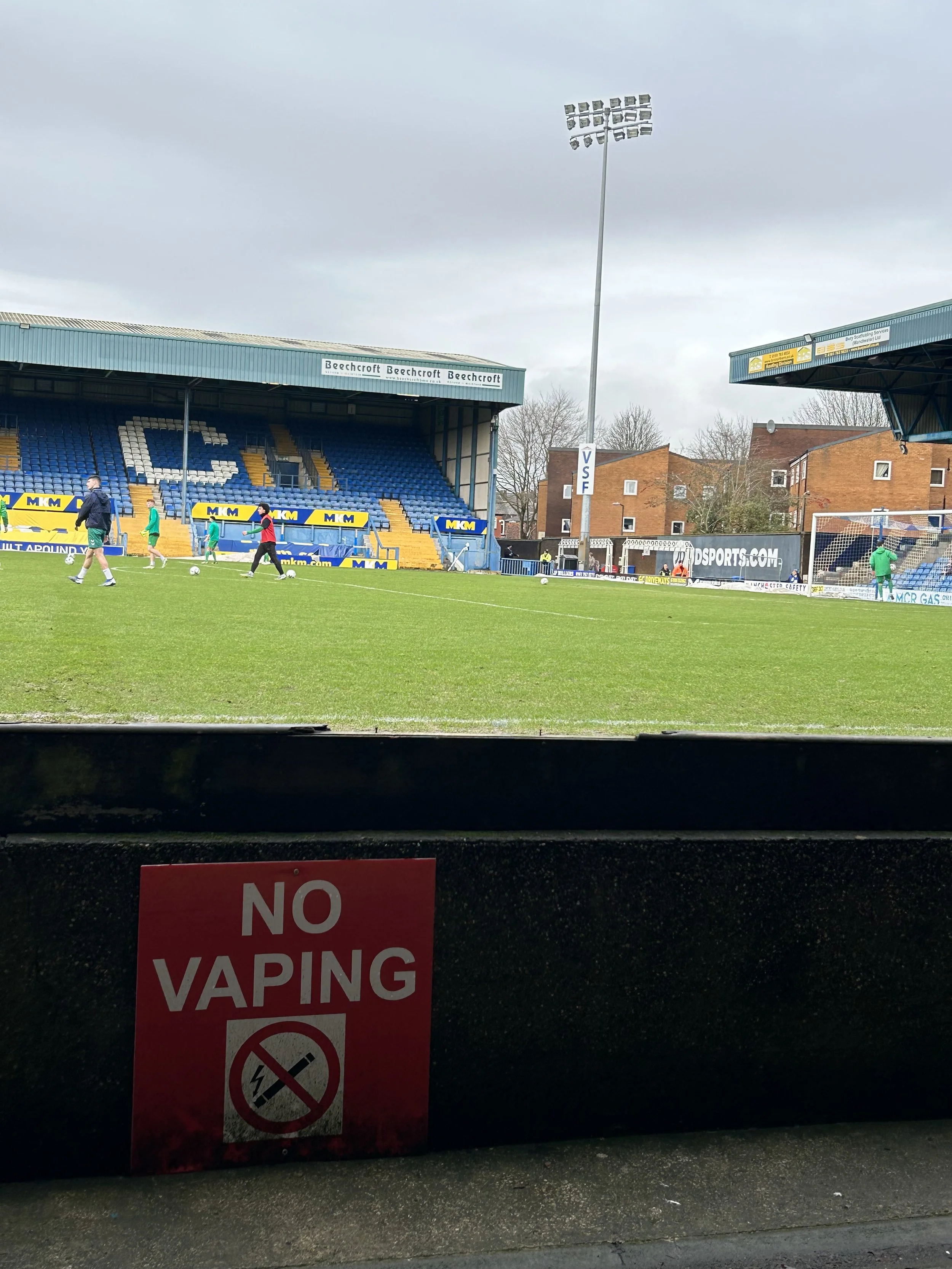 View of a soccer stadium with players warming up on the field, stadium seating, and a sign that says 'NO VAPING' in the foreground.