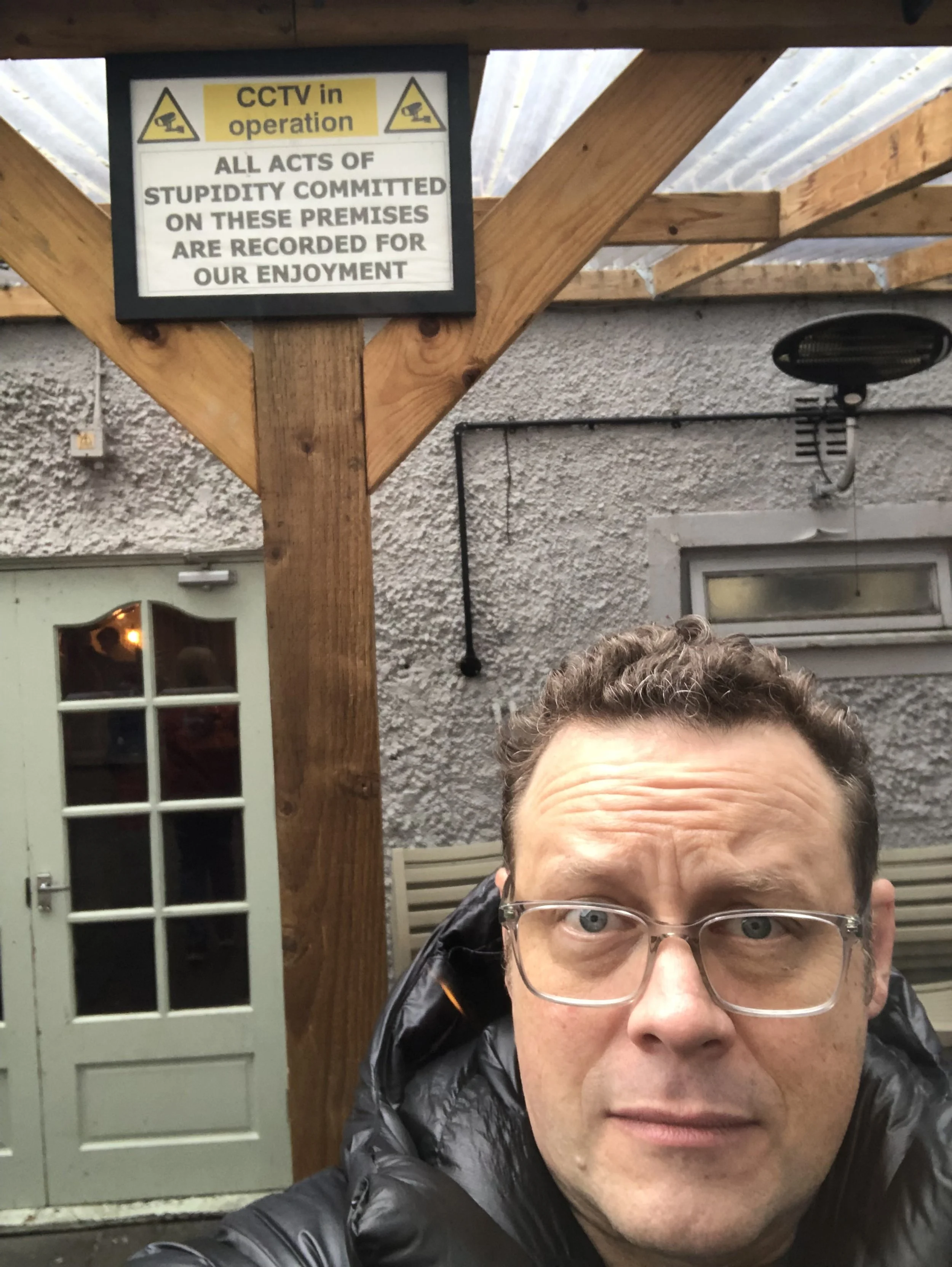 A man taking a selfie outdoors with a gray, textured wall, a green door with windows, and a wooden structure with a CCTV sign in the background.