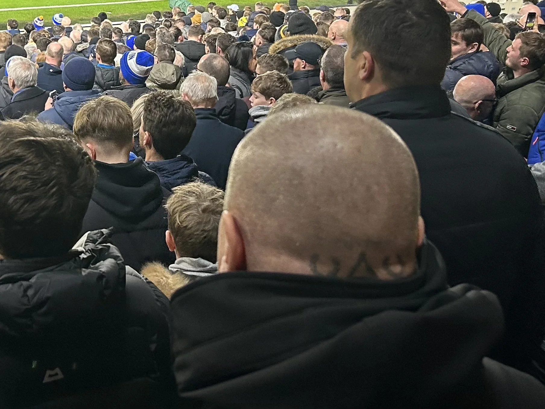 Crowd of people at a sporting event, seen from behind, with many wearing dark clothing and some wearing hats or beanies.