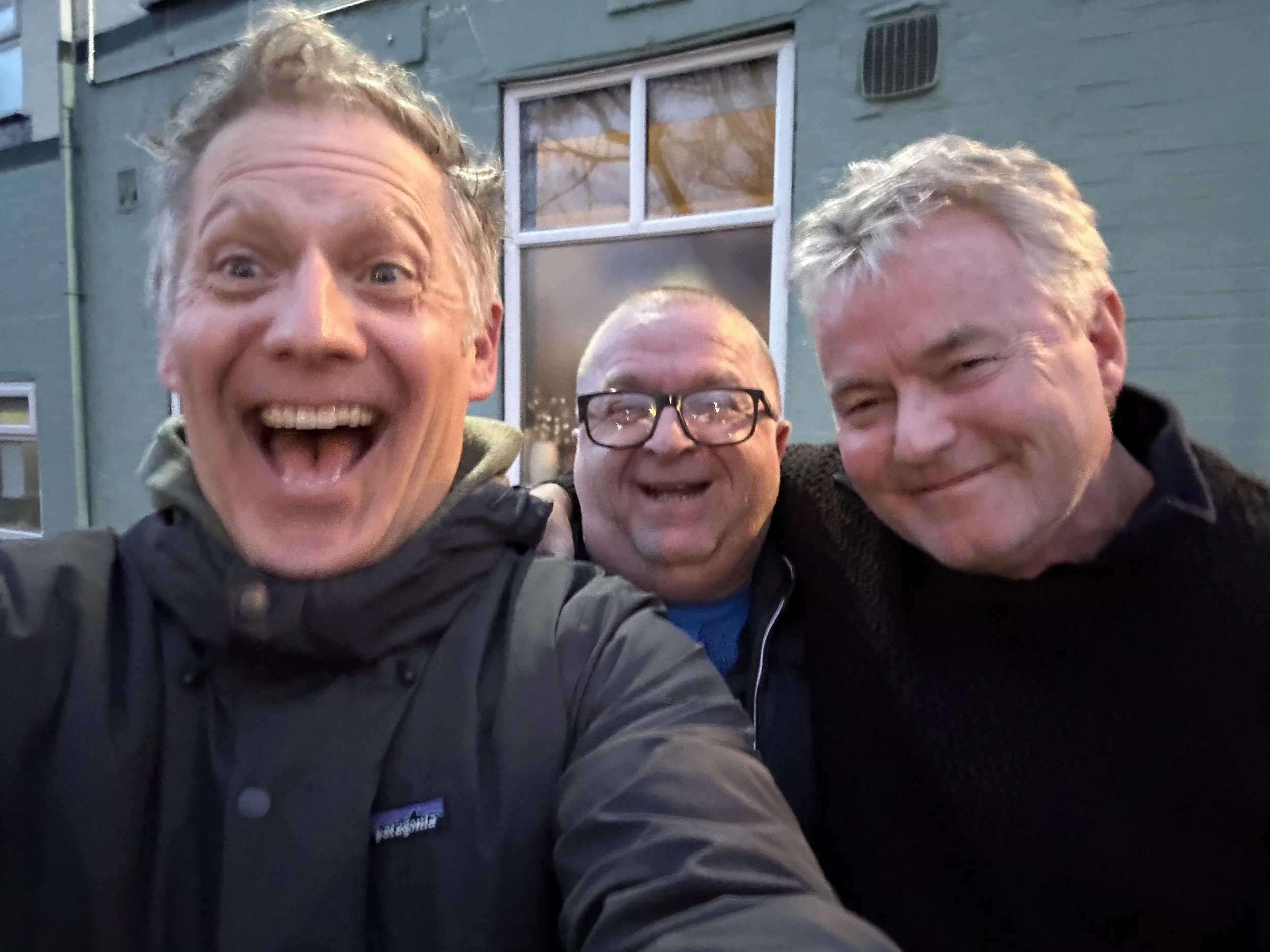 Three middle-aged men smiling and posing for a photo outdoors at dusk, with a house and window in the background.