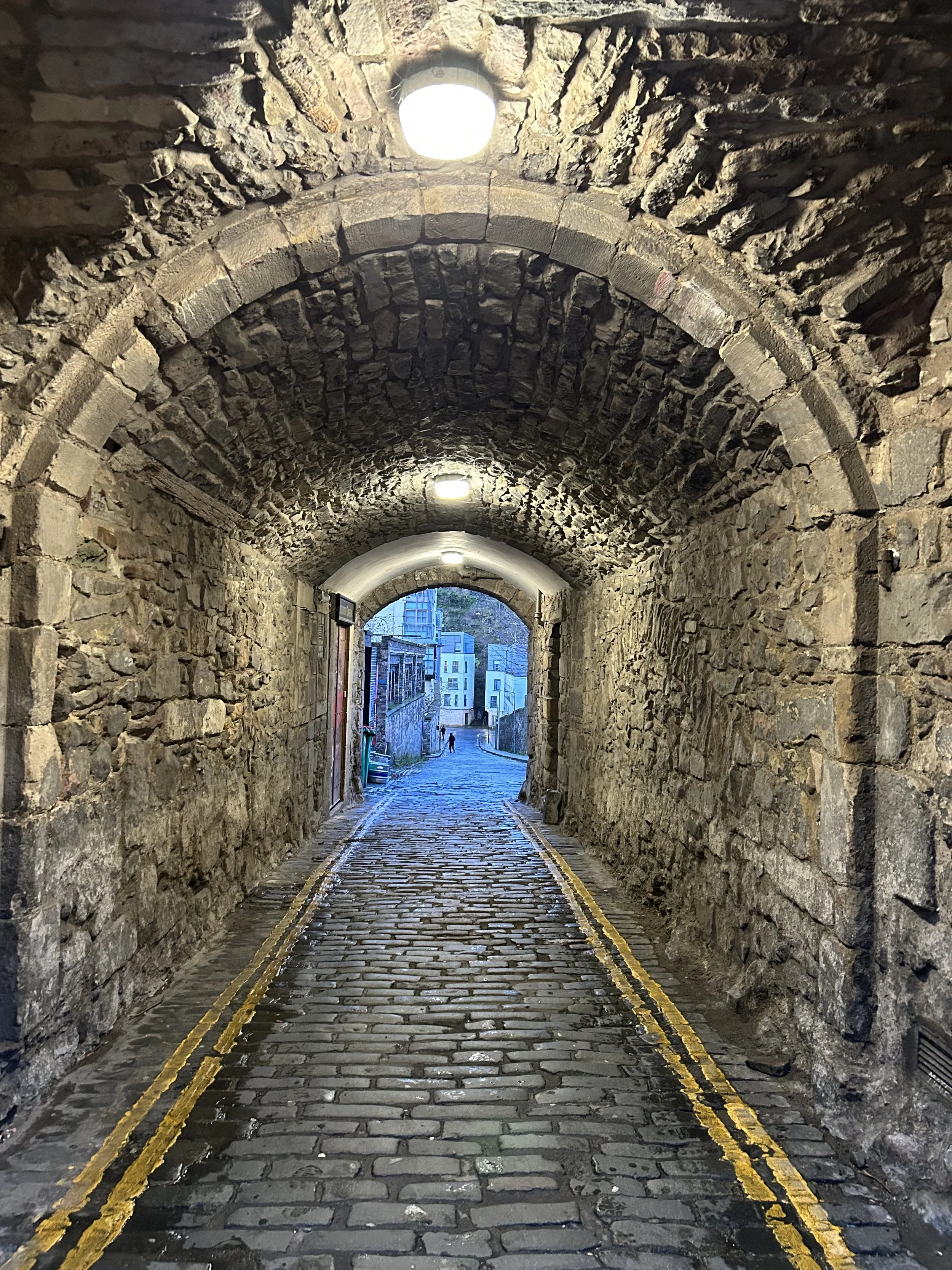 A cobblestone alleyway viewed from under a stone arch tunnel with brick walls and yellow lines along the edges, leading to a street with modern buildings and a person walking in the distance.