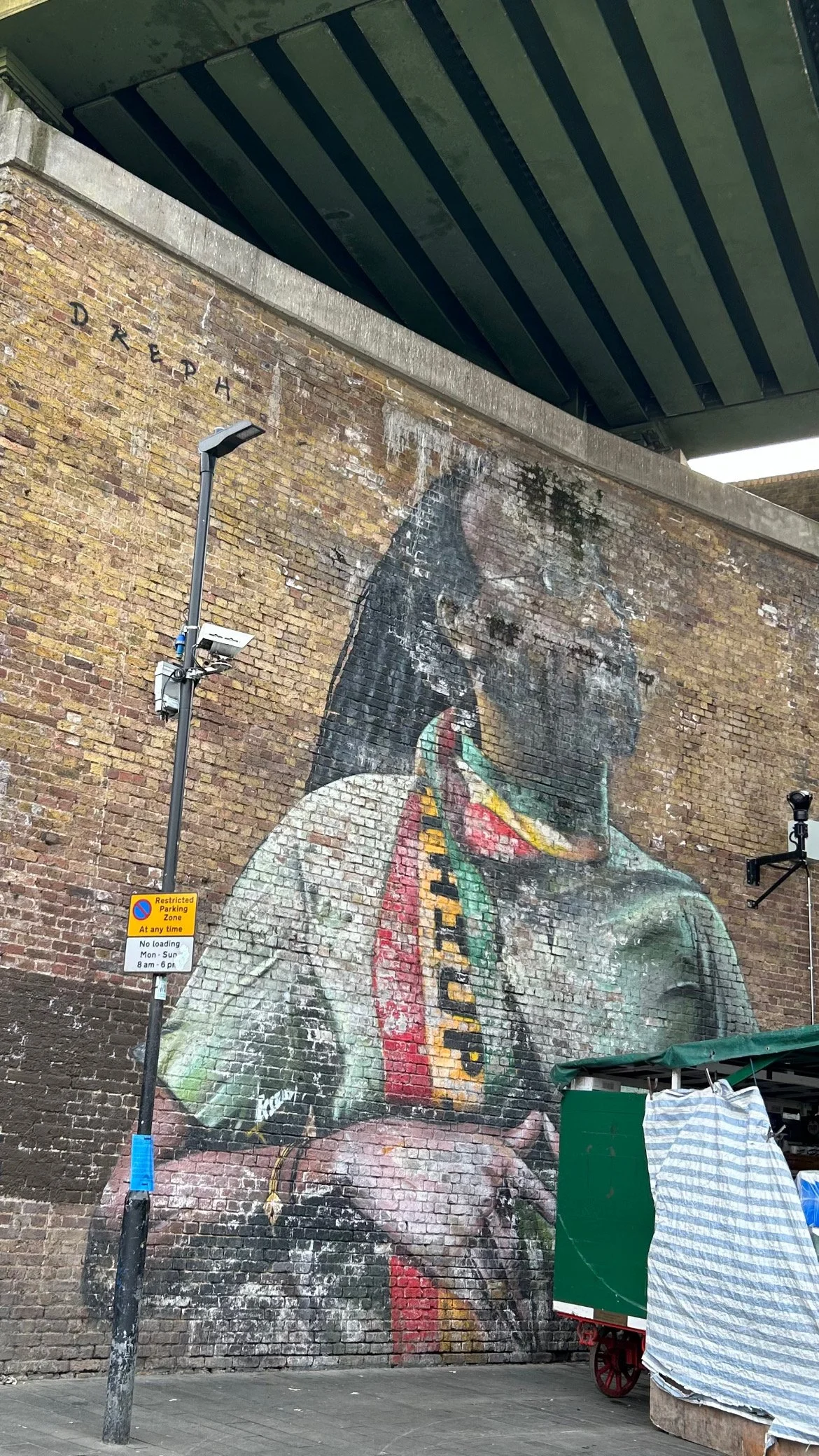 Large mural of a person with dreadlocks and dark skin, wearing a colorful scarf and holding their hands together, painted on a brick wall under a bridge.