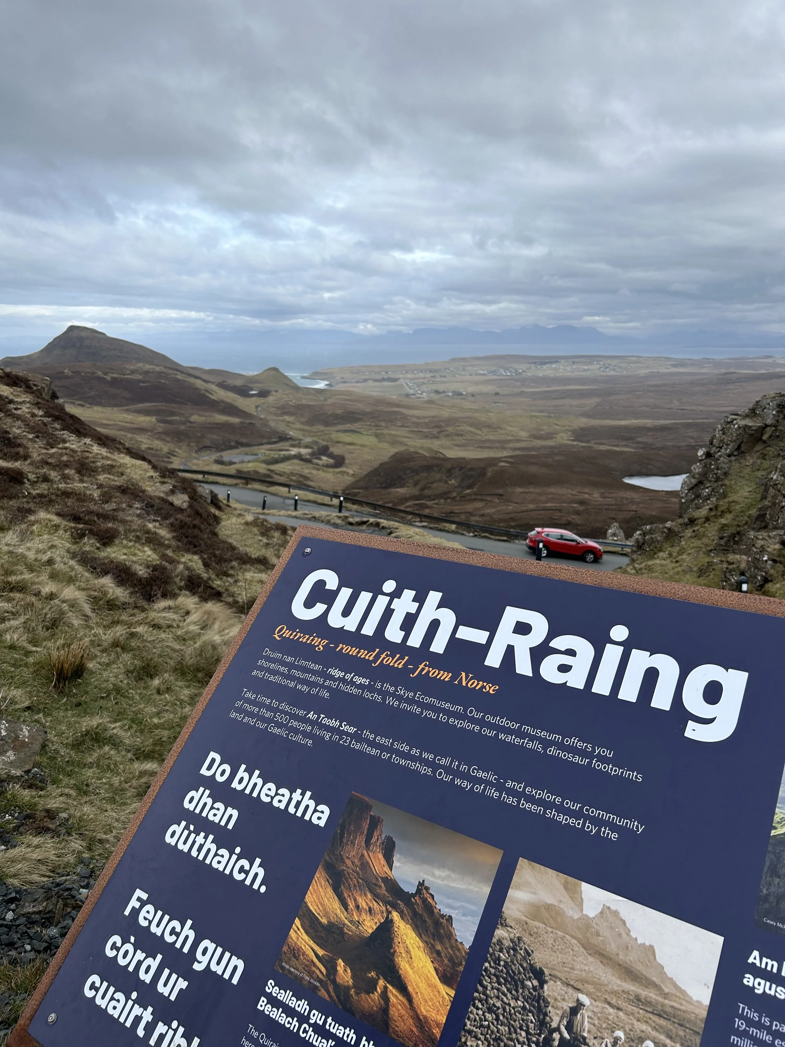 View of a mountainous landscape with a road winding through the terrain, under a cloudy sky. There is an informational sign about Cuith-Raing in the foreground.