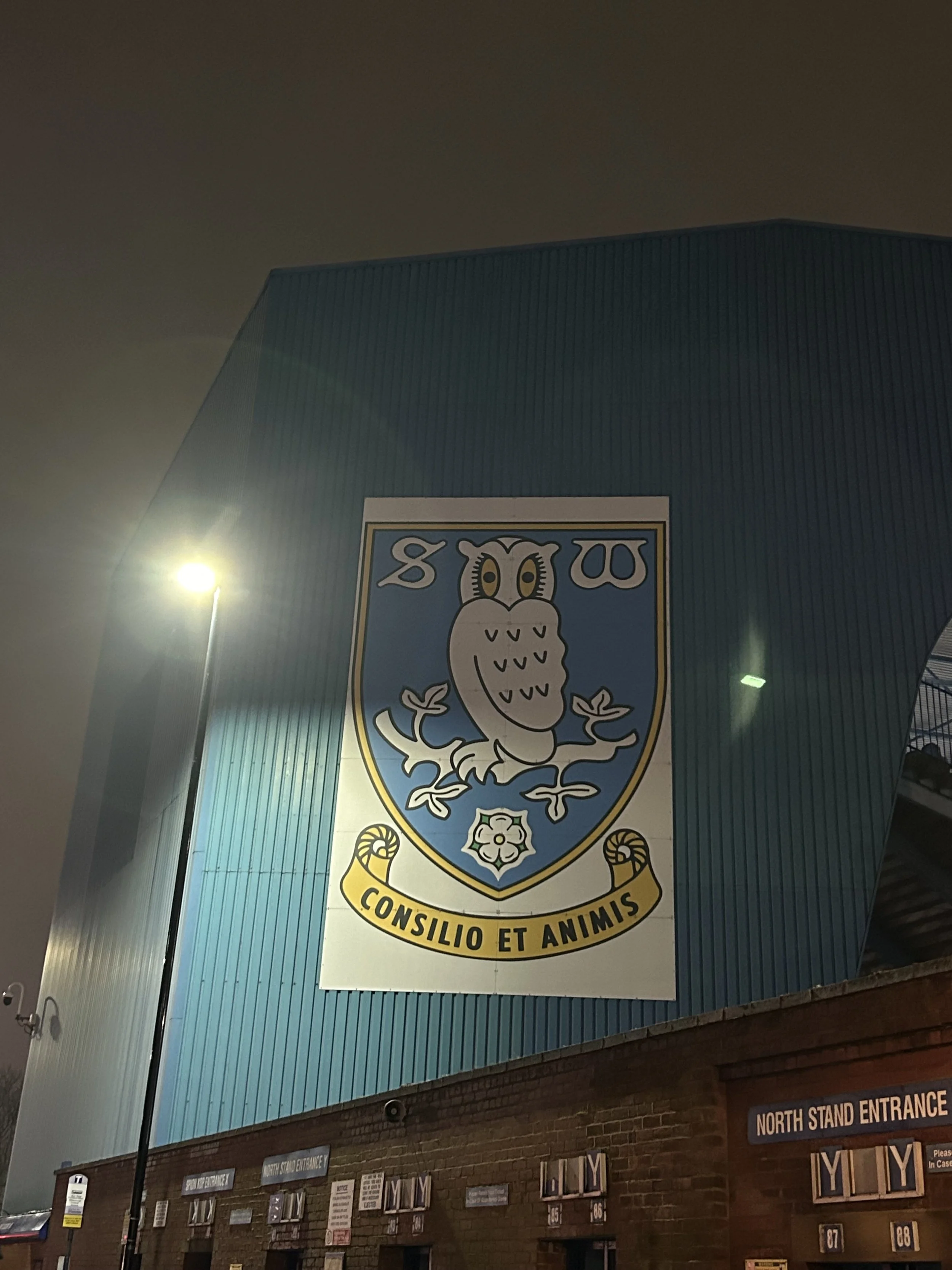 Large crest with an owl sitting on a branch, the crest has a blue background with a white border, and the words 'CONSILIO ET ANIMIS' at the bottom. The building is blue with a brick base and is labeled 'North Stand Entrance'.