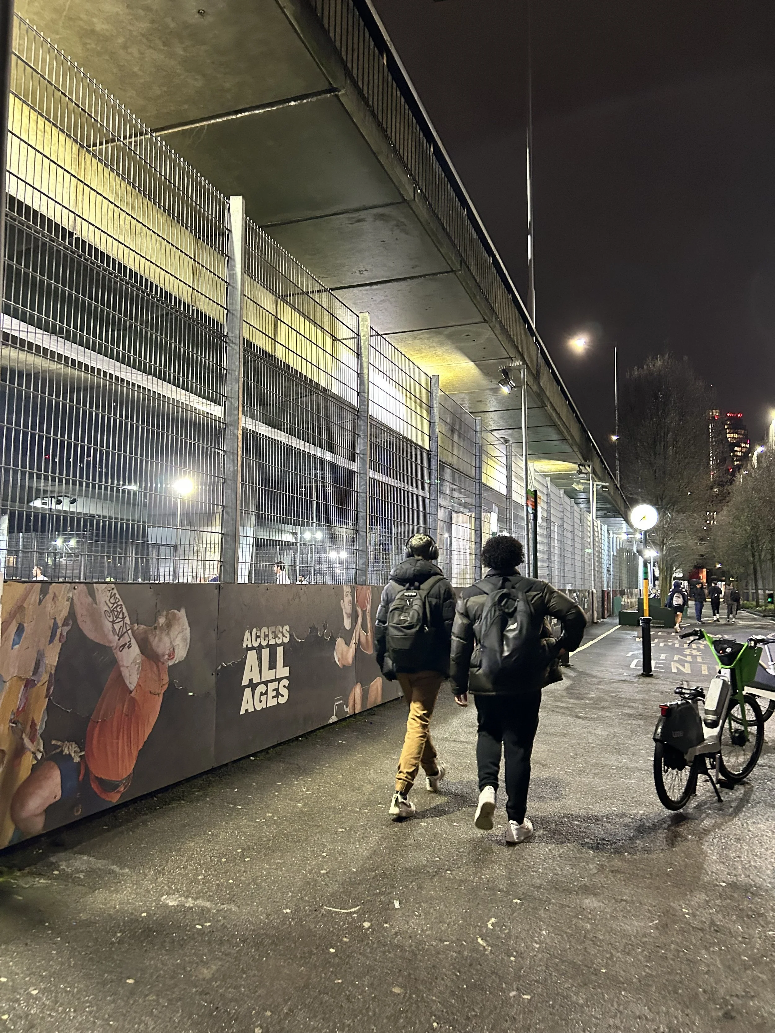 Two people walking on a city sidewalk at night, near a fenced sports court with a sign that reads 'ACCESS ALL AGES'; bicycles are parked nearby and streetlights illuminate the scene.