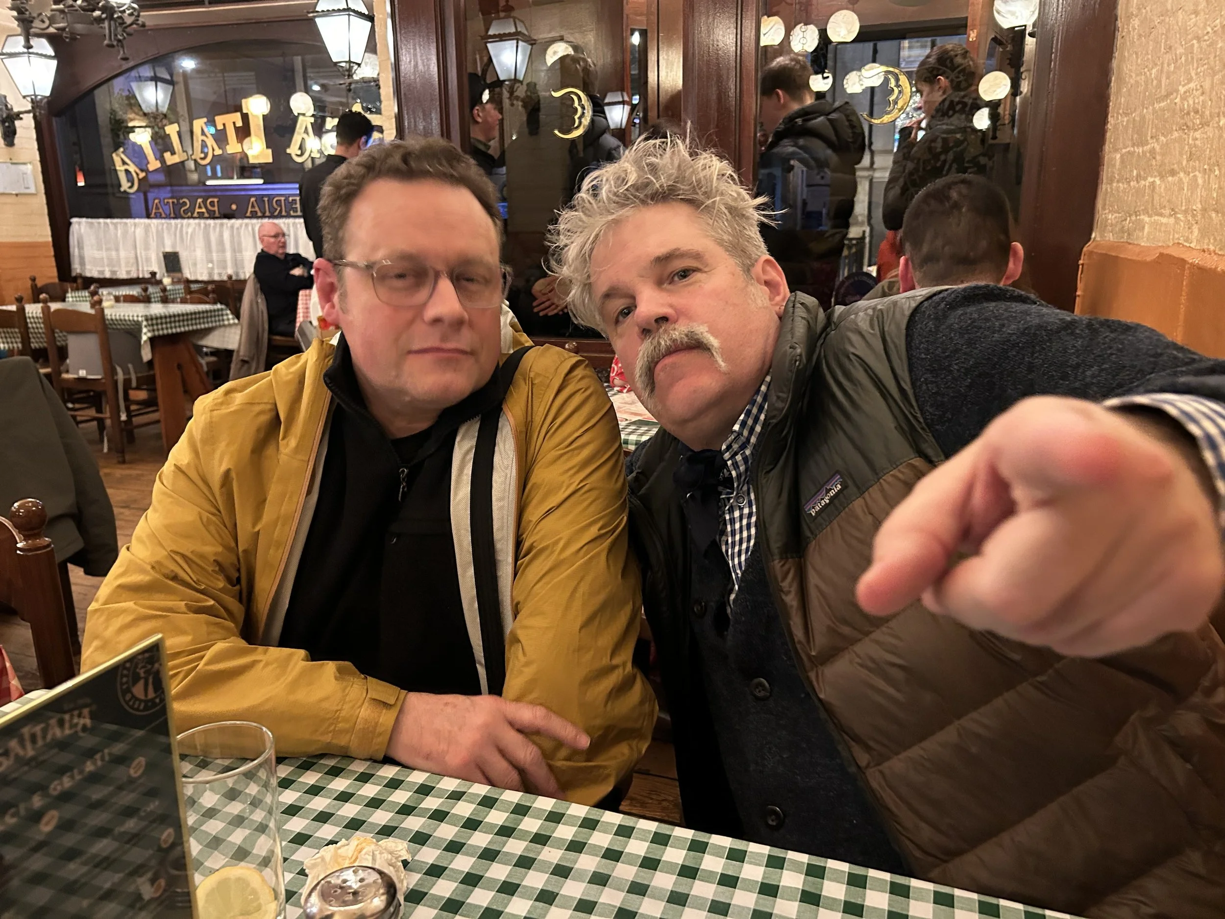 Two middle-aged men sitting at a restaurant table with a checkered tablecloth, looking at the camera. The man on the left is wearing glasses and a yellow jacket, while the man on the right has a mustache, messy gray hair, and is pointing toward the c