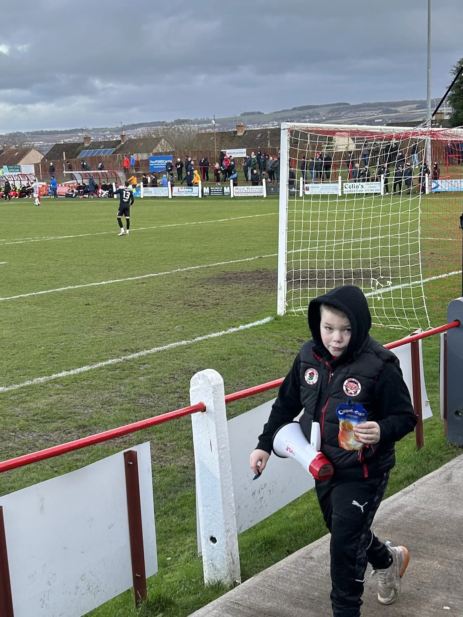 A young boy dressed in a black hoodie and sports jacket holding a drinks carton and a megaphone stands near the fence at a soccer match, with players and spectators in the background on a cloudy day.