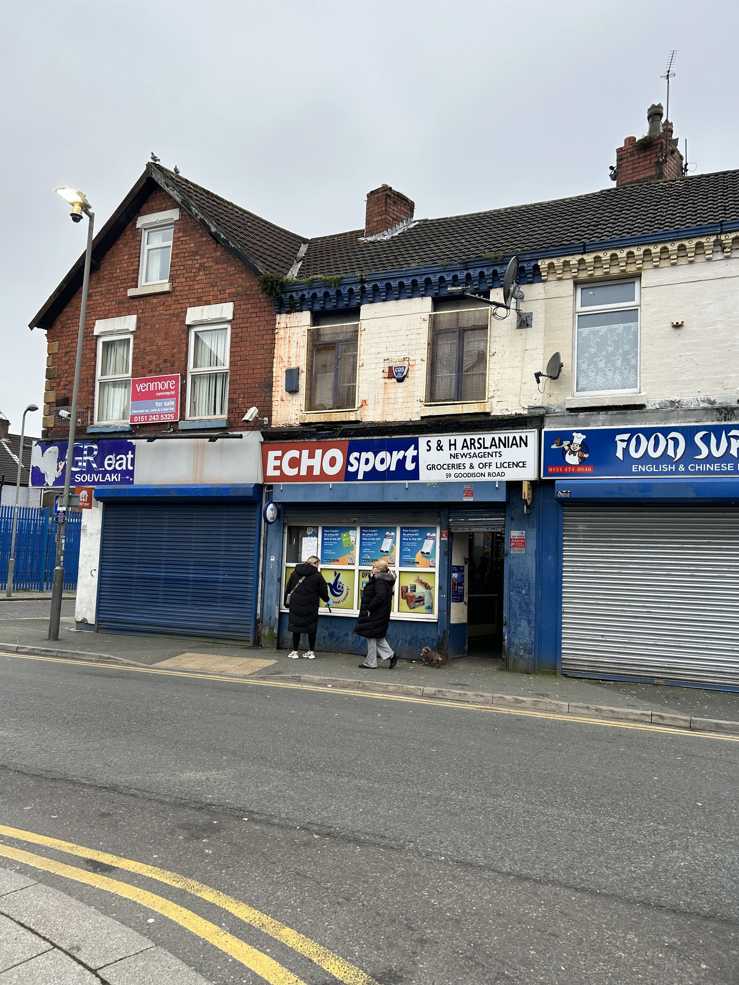 Street scene with two women in winter jackets talking outside a small store named ECHO sport, with a blue and red sign, and neighboring shops with shutters down. There is a building above with damaged graffiti-covered windows.