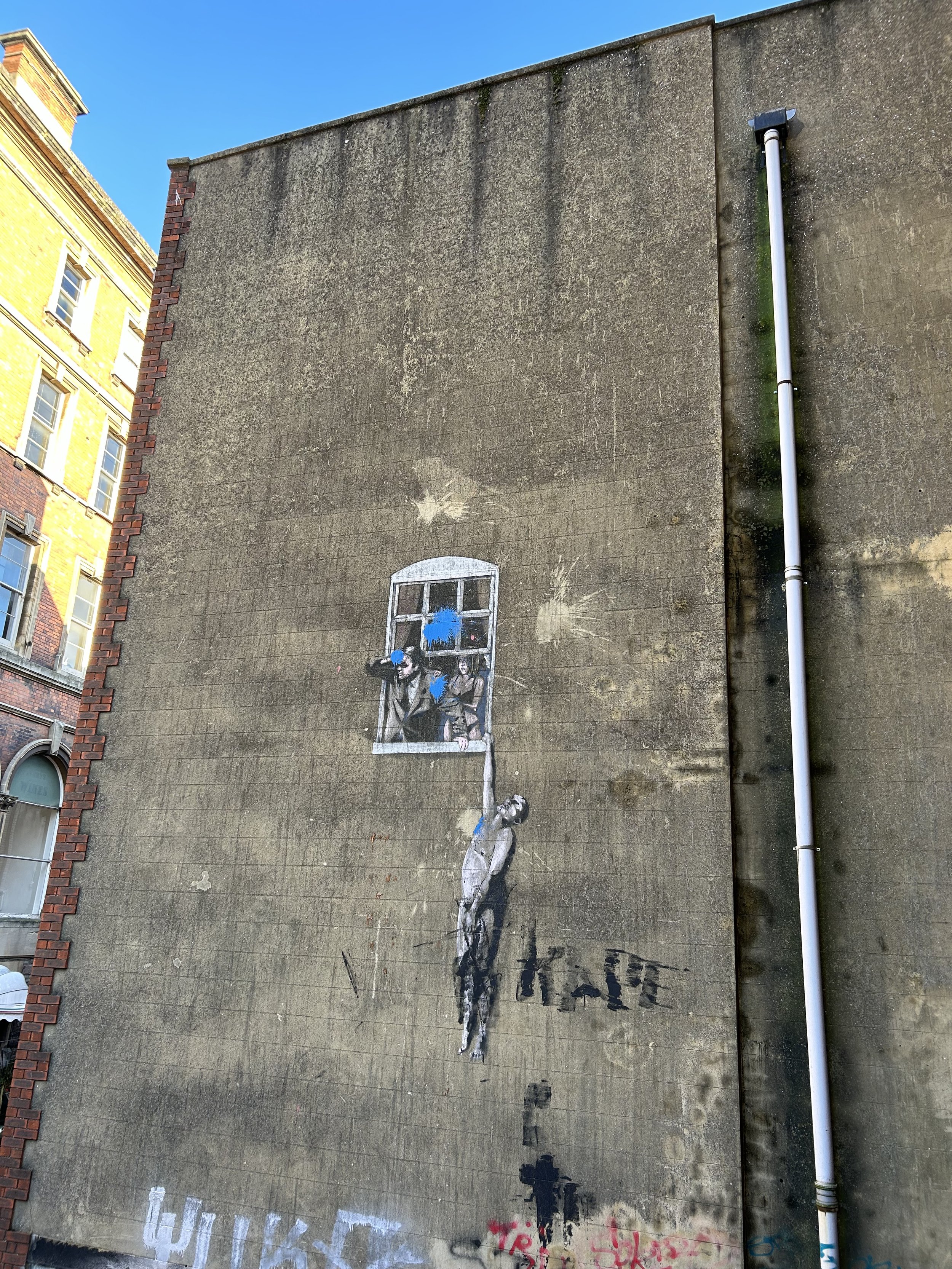 Street art mural on a concrete wall depicting a woman standing beneath a window with two men leaning out of the window, one holding a blue paint spray. The woman is connected to the window by a blue line. The wall has various graffiti tags and paint 