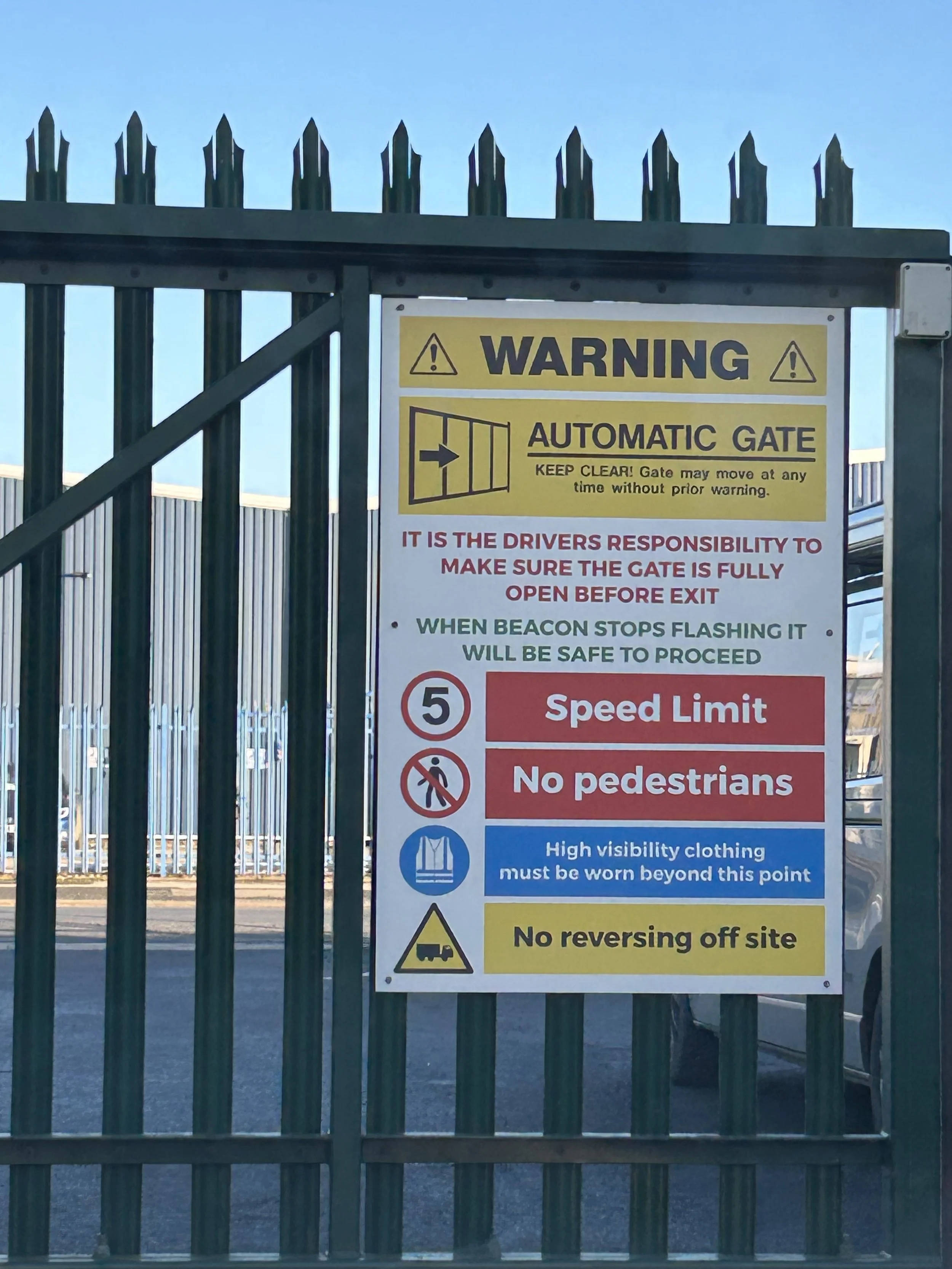 Sign on a green gate with instructions about an automatic gate, including speed limit, no pedestrians, safety clothing requirement, and no reversing, with a background of metal fencing and vehicles.