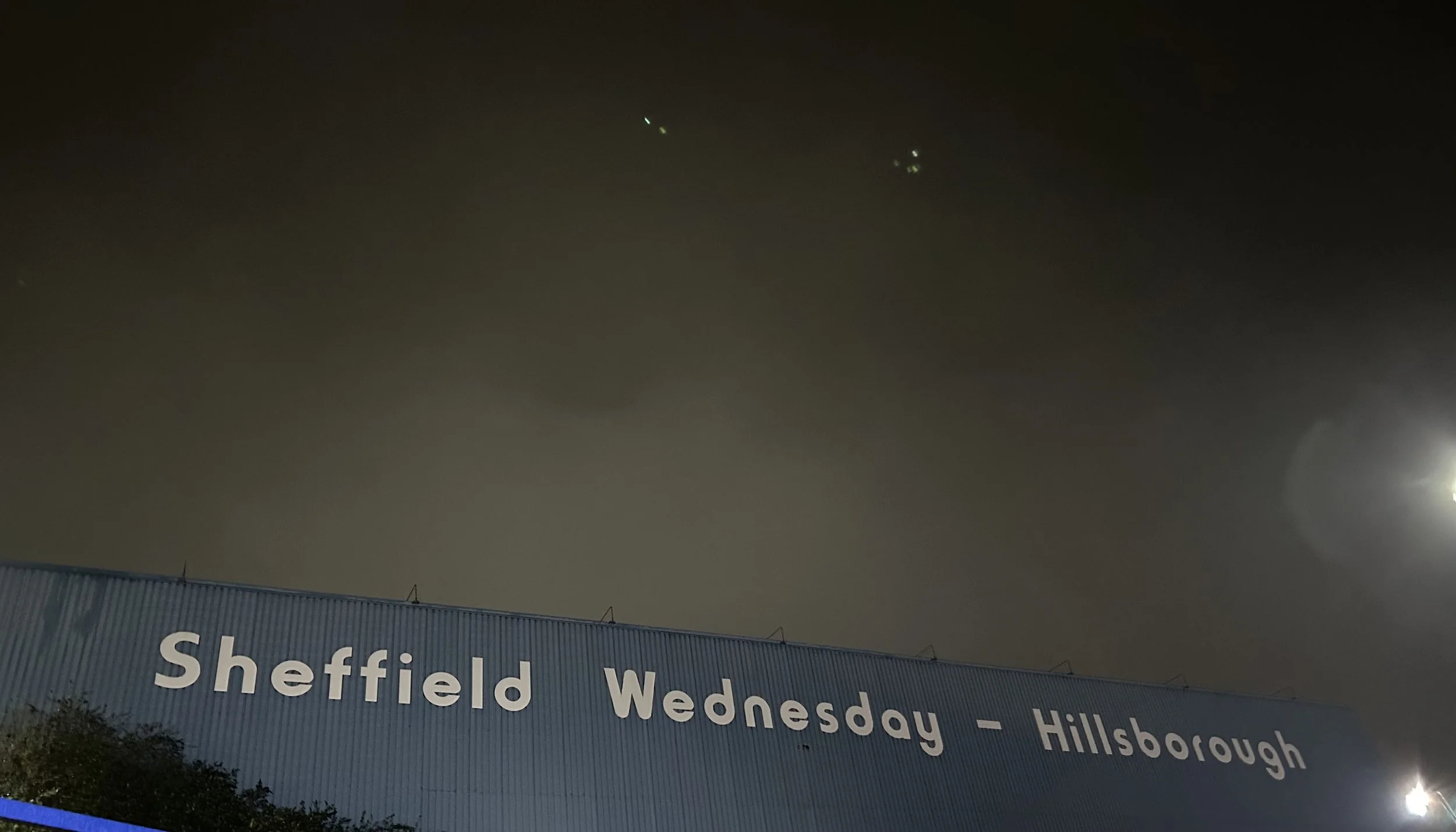 Nighttime view of a large blue sign with white text reading 'Sheffield Wednesday - Hillsborough' under a dark sky with a bright moon and some faint stars.