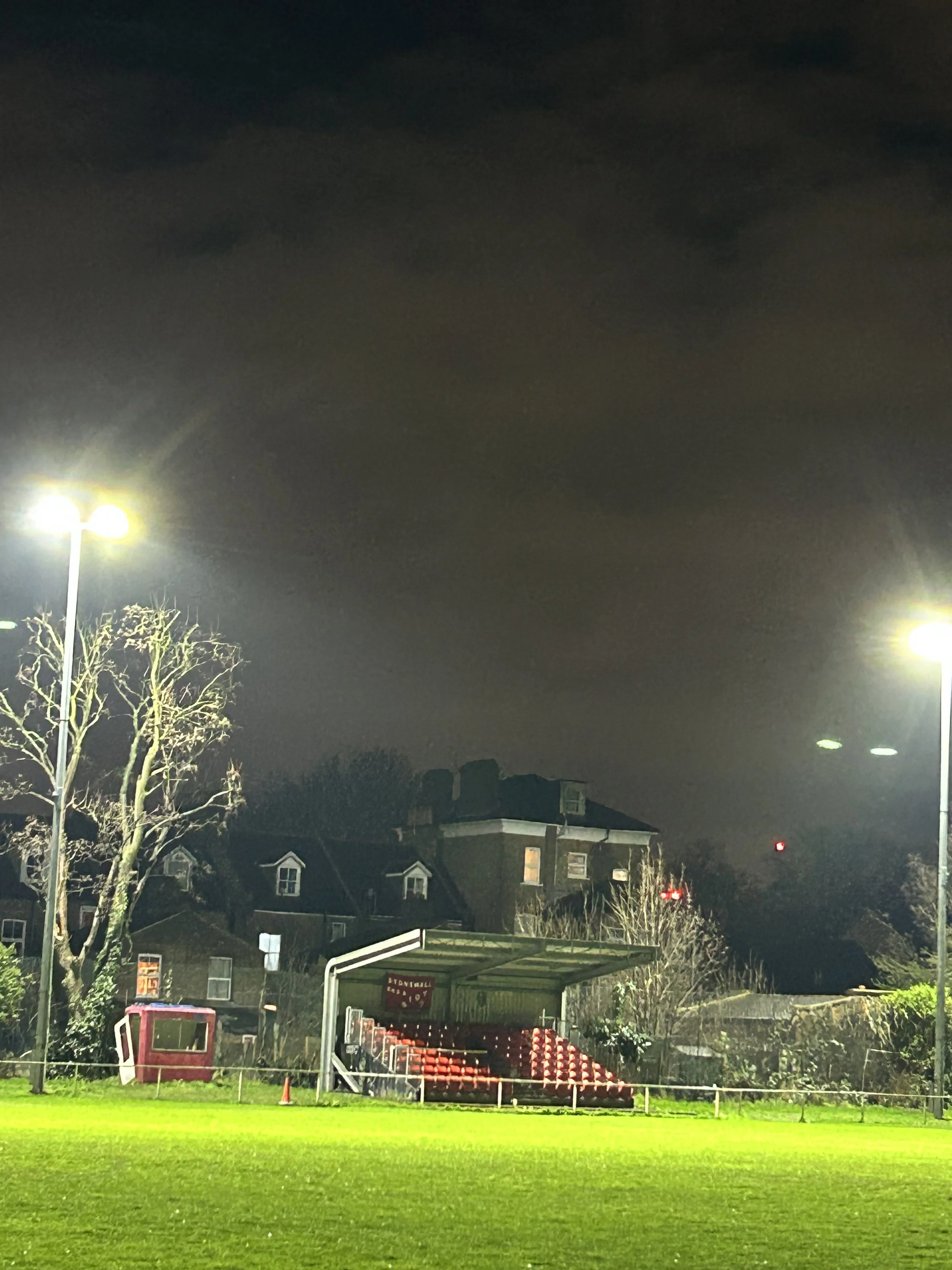 Nighttime view of an empty sports stadium with bright floodlights, green grass field, and red seating area with a small covered section, located near residential buildings.