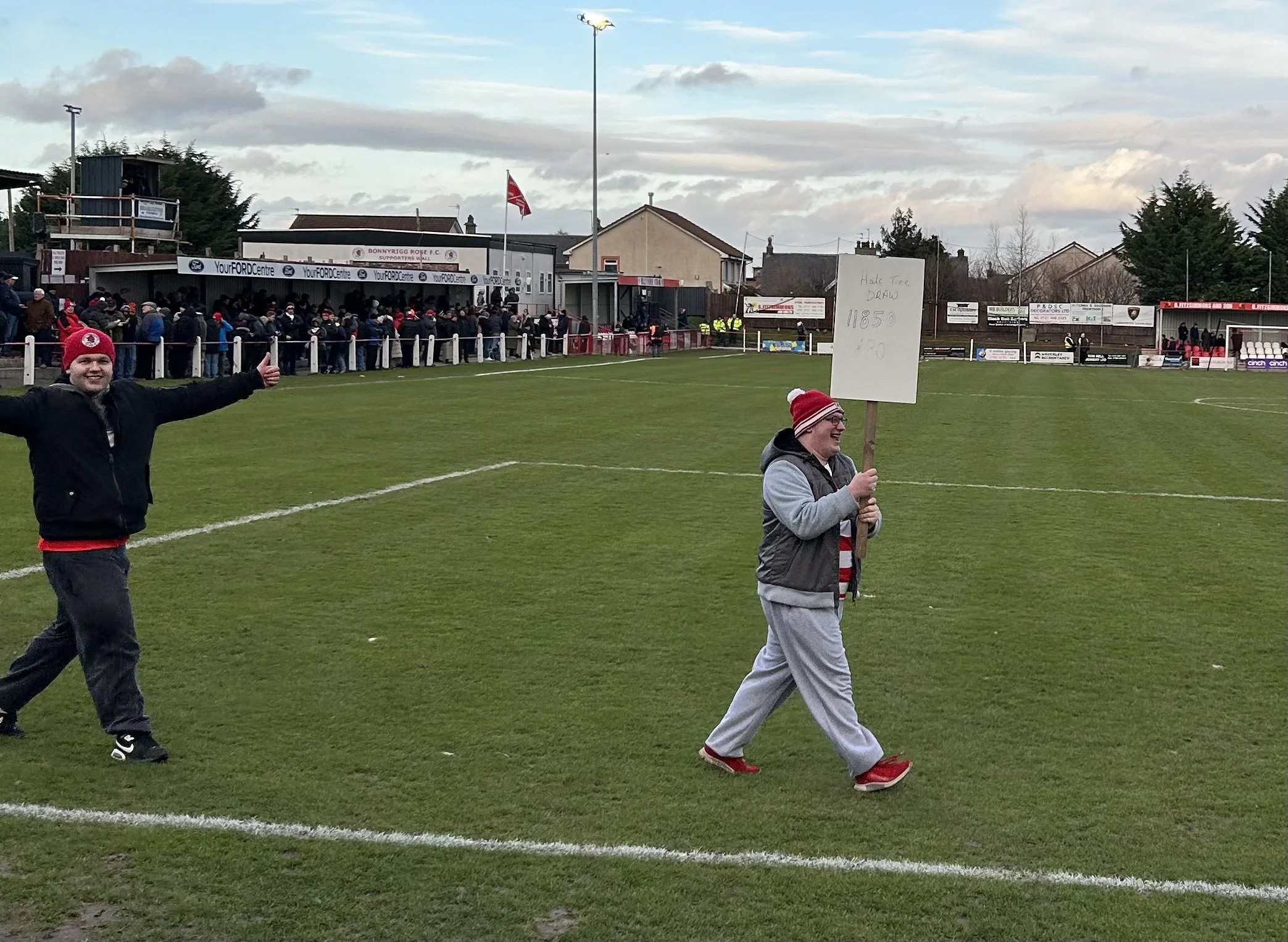 A person holding a sign walking onto a soccer field, with another person giving a thumbs-up nearby. Spectators and a stadium are visible in the background, and the person with the sign is wearing a red and white hat.