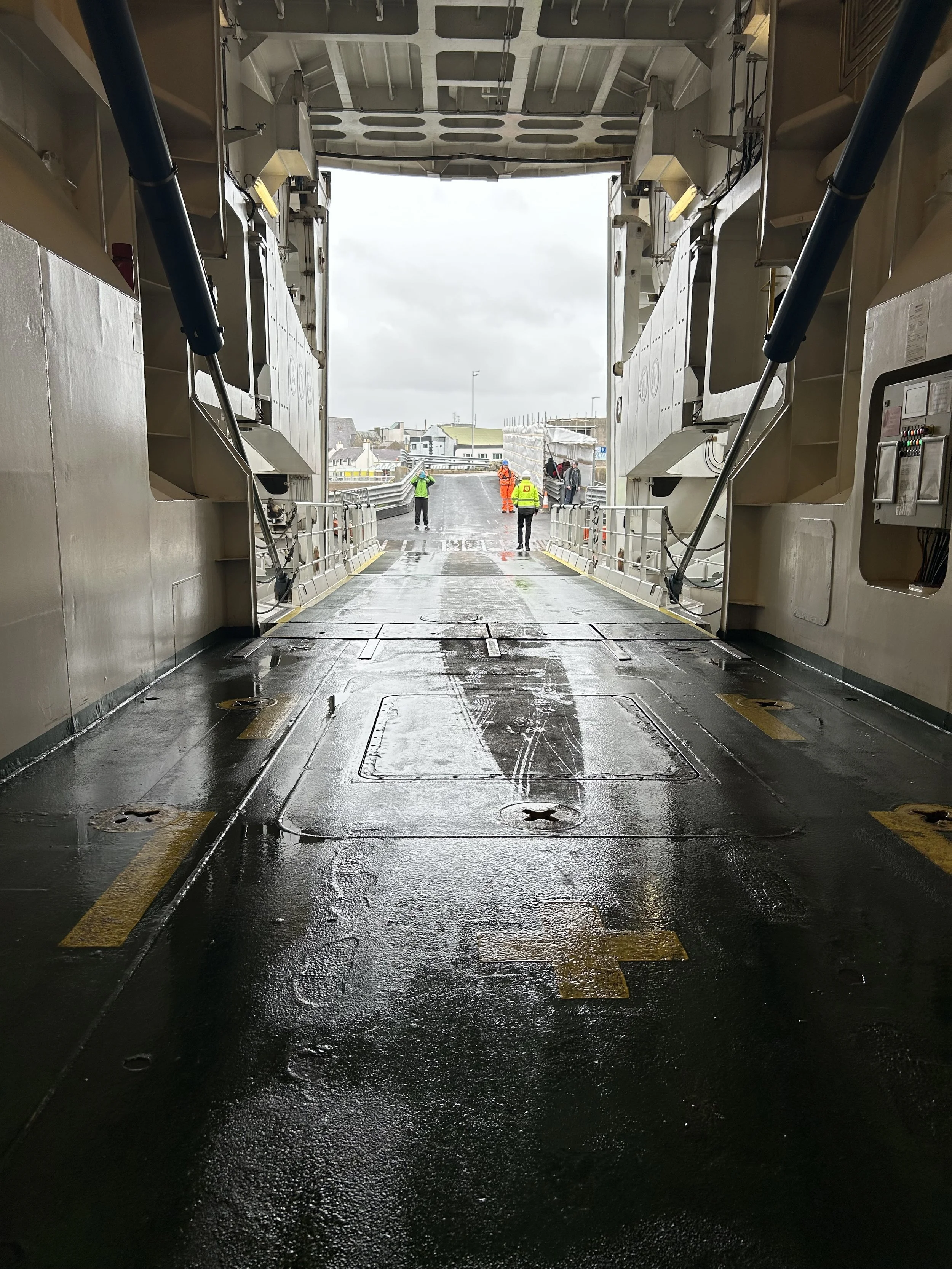 The image shows the interior of a large, open cargo ship or aircraft with a wet, black metal floor and several anchor points. Outside, on the dock, there are four workers wearing high visibility clothing, some in orange and some in green, engaged in 