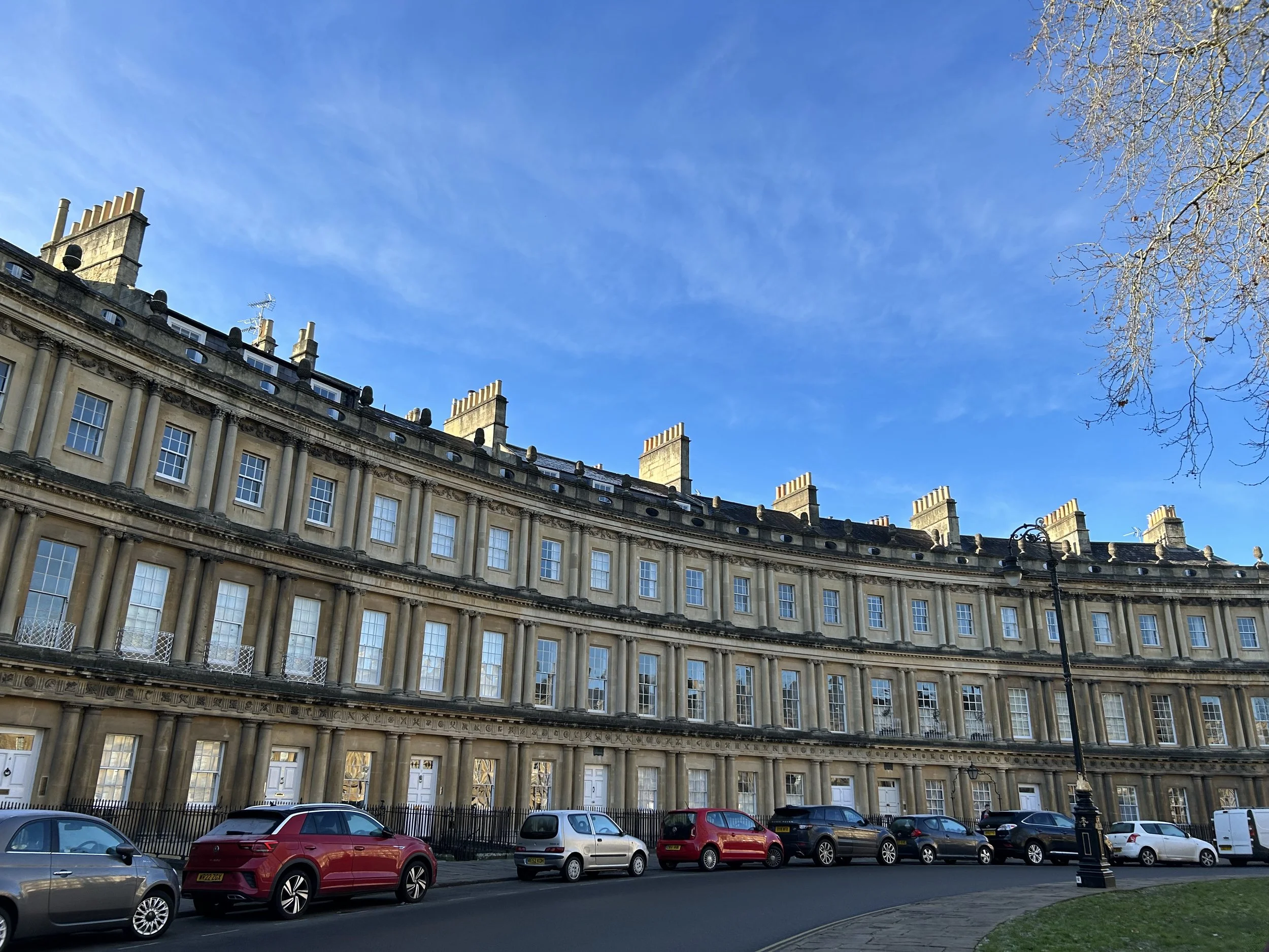 A curved row of historic beige stone buildings with numerous windows and chimneys, parked cars along the street, a decorative black lamppost, a leafless tree, and a clear blue sky.