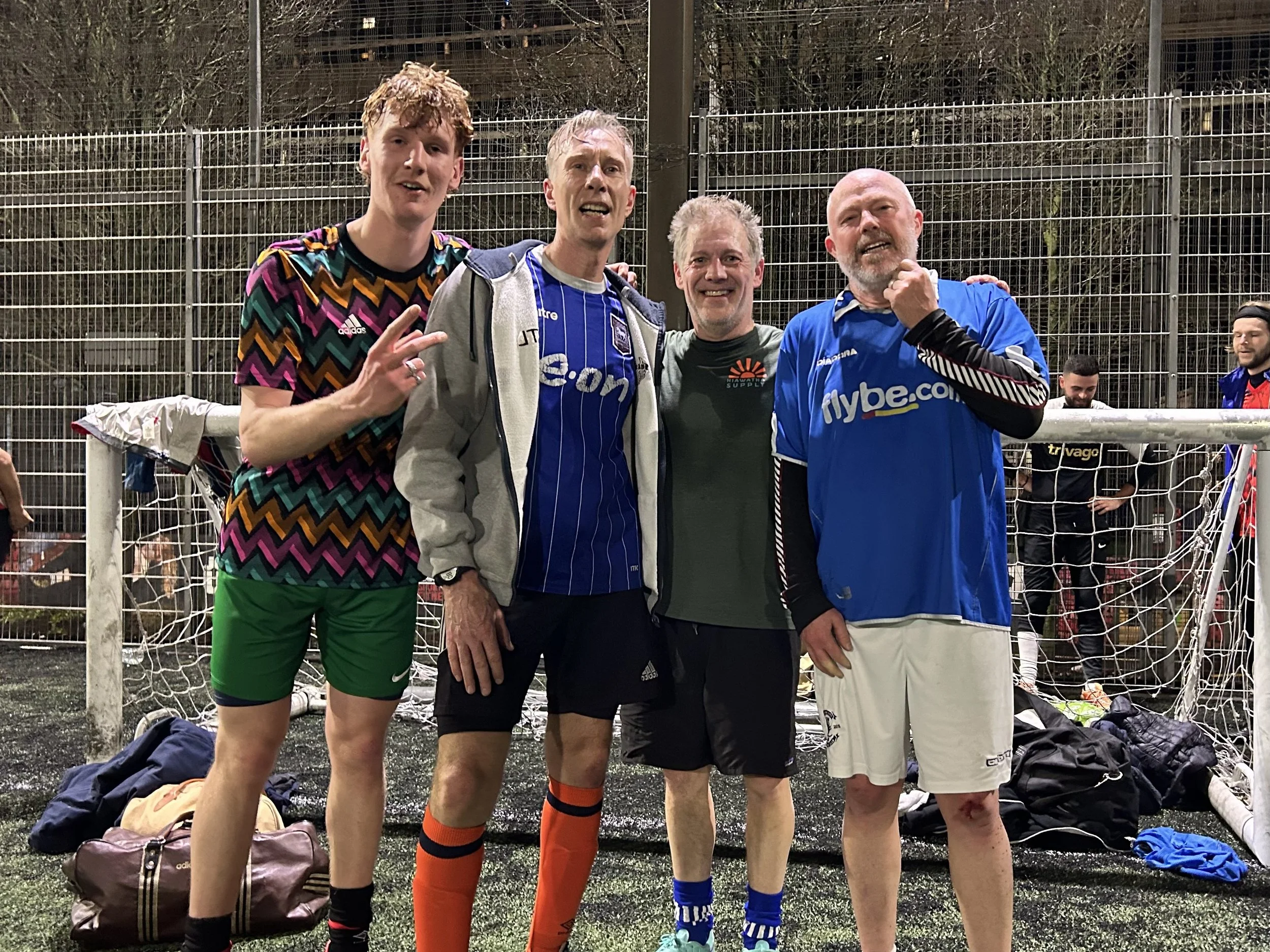 Four men standing together on a soccer field at night, smiling, with a goal net behind them, after playing soccer.
