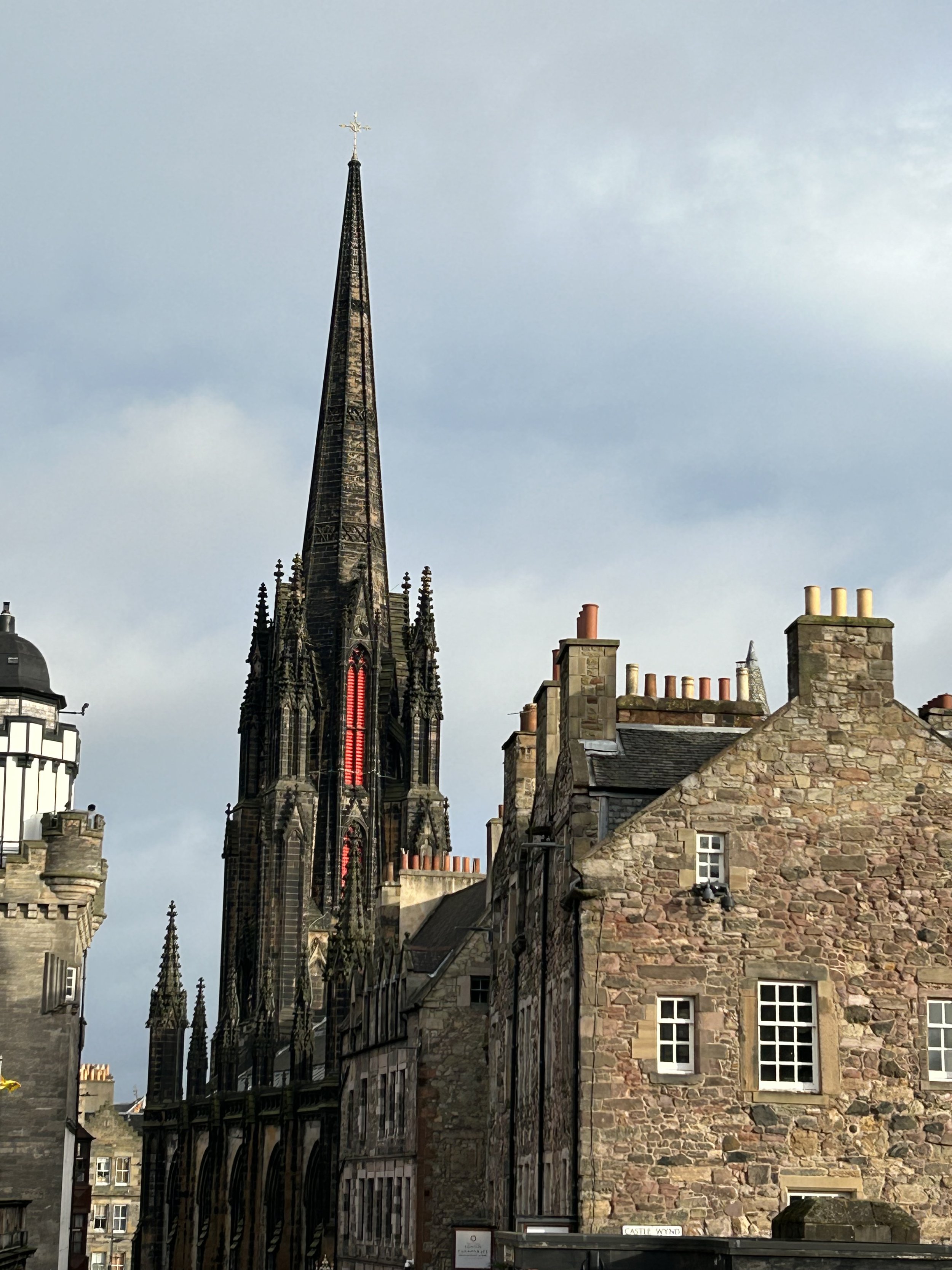A Gothic-style church with a tall spire and a cross at the top, situated behind old stone buildings with chimneys, under a cloudy sky.