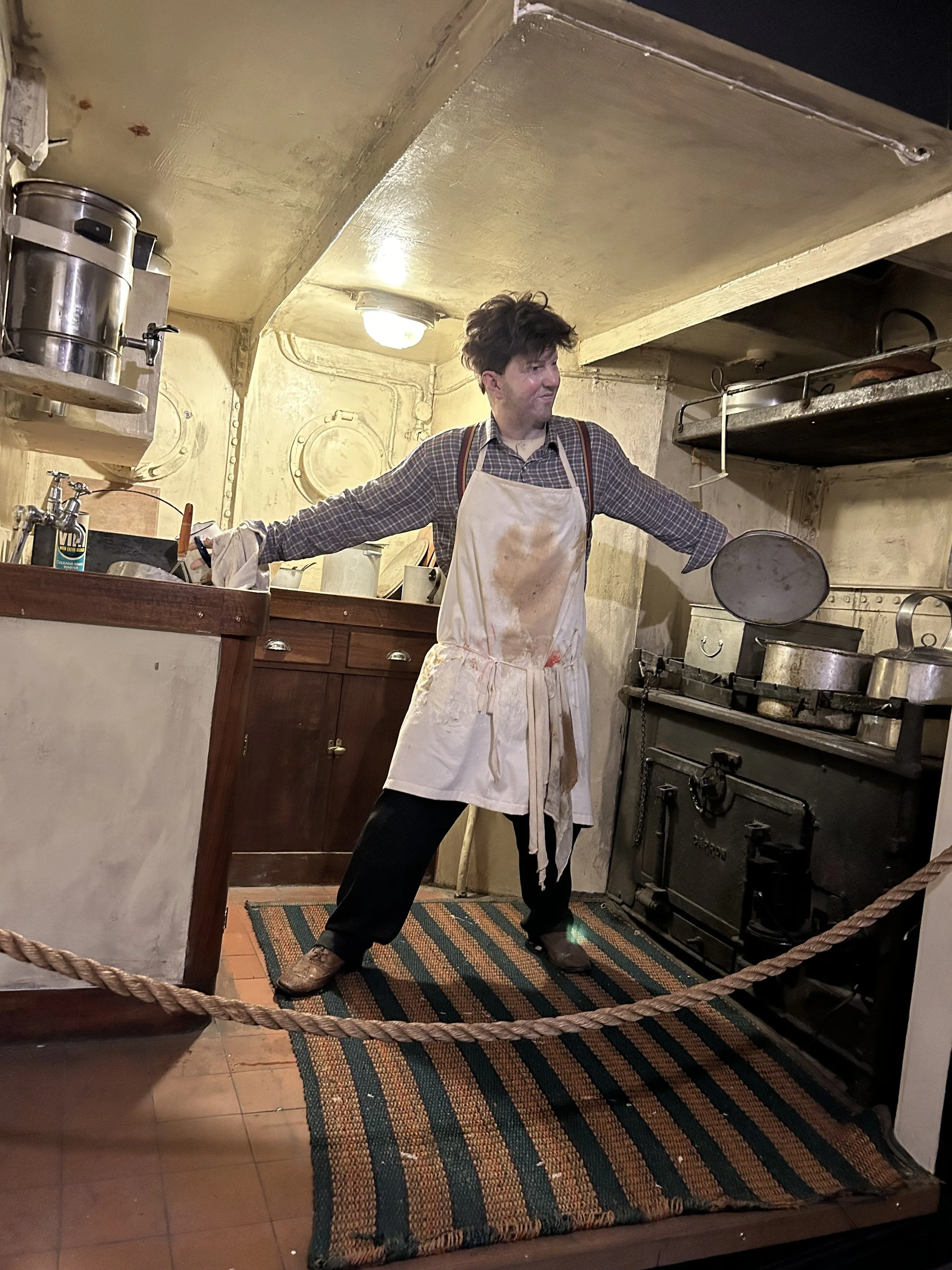 Man in a kitchen dressed as a cook, wearing a stained apron, holding a lid, with an old stove and pots in the background.