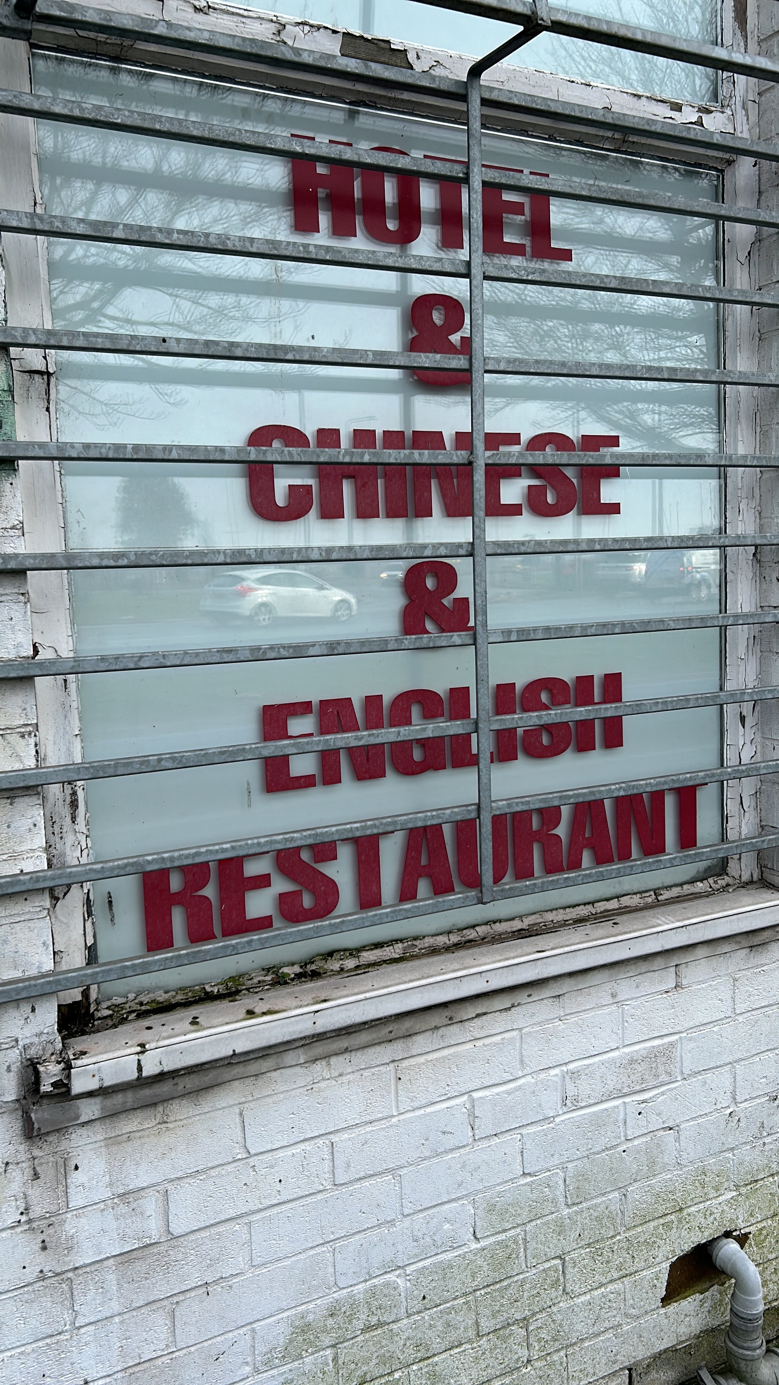 Sign reading 'Hotel & Chinese & English Restaurant' behind metal bars, on a white brick wall with a window and a pipe below.
