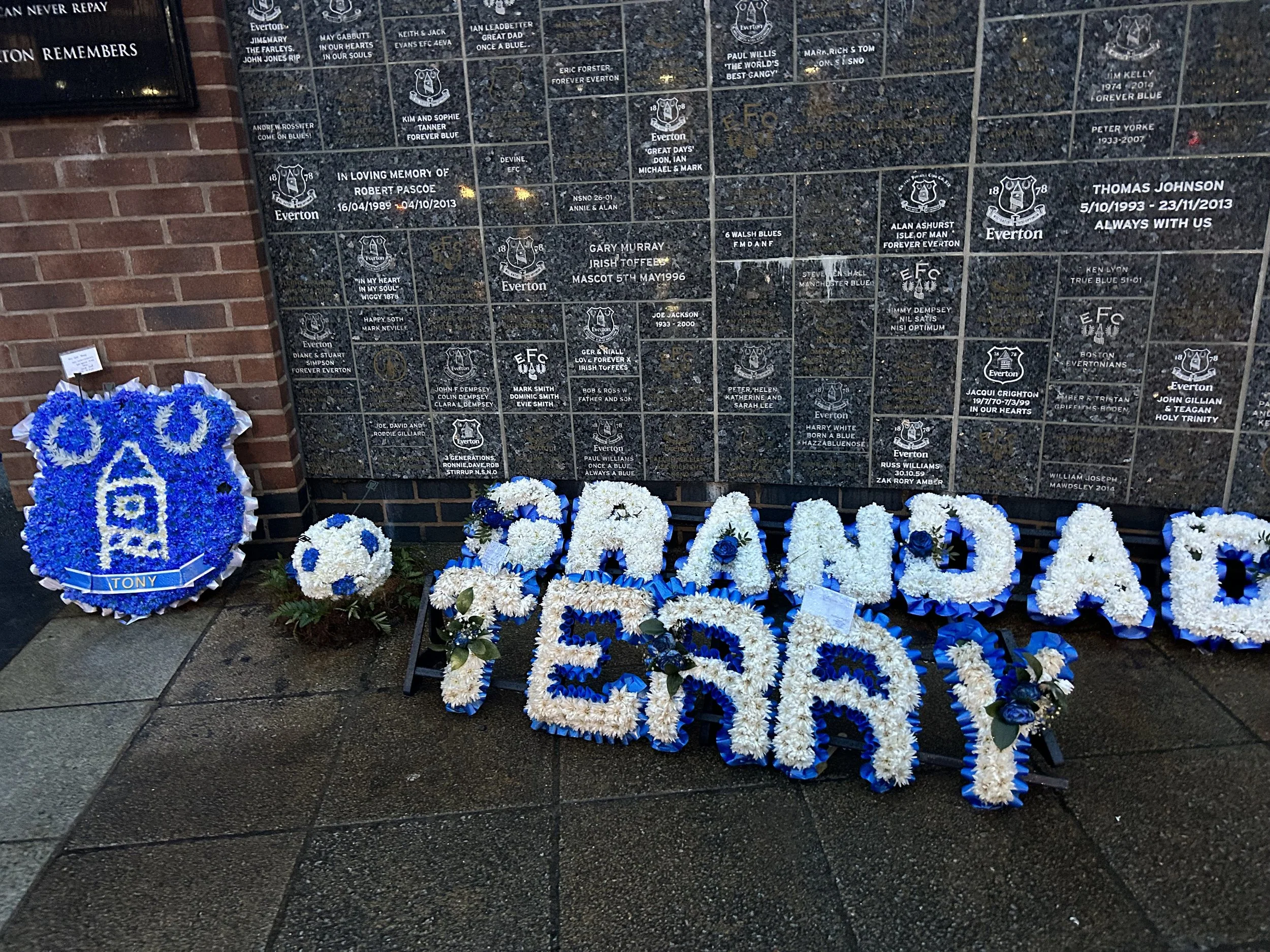 Memorial wall with floral tributes: large blue and white flower arrangement with a church emblem labeled 'Tony', and word 'EERA' in large white and blue flowers, with smaller flower arrangements and a plaque with names and dates.