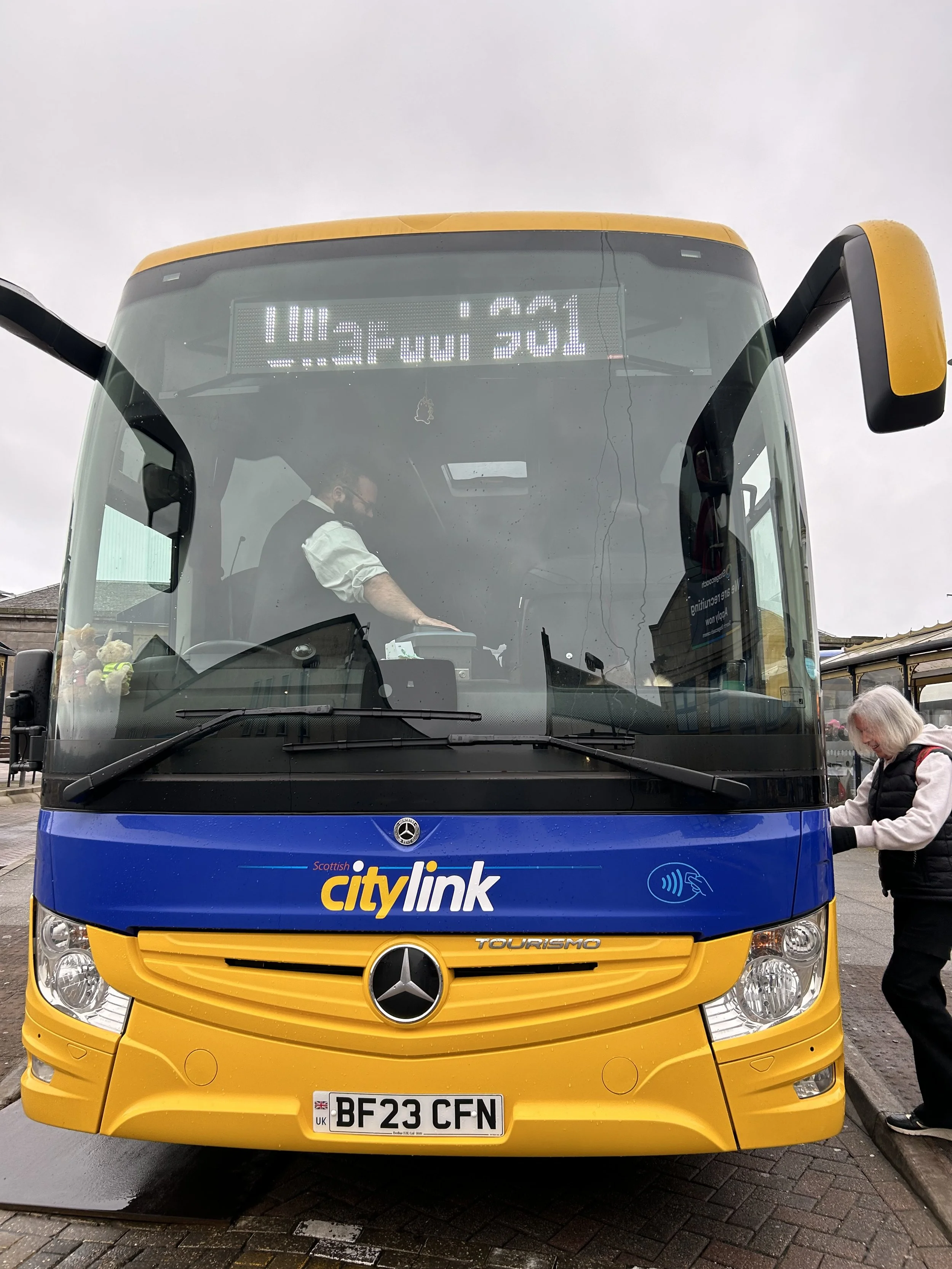 Front view of a city bus with a blue and yellow color scheme, displaying a sign with route information and a woman boarding on the right side.