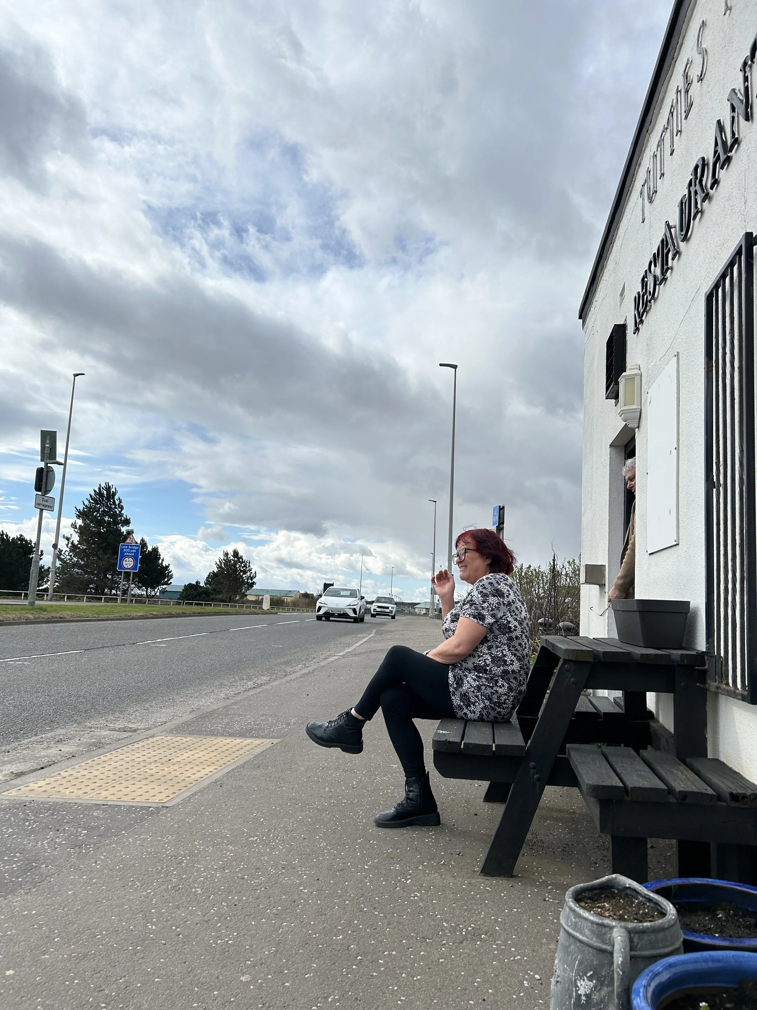 A woman with red hair and glasses sitting on a bench on the sidewalk outside a white building, with cars on the road and cloudy sky in the background.