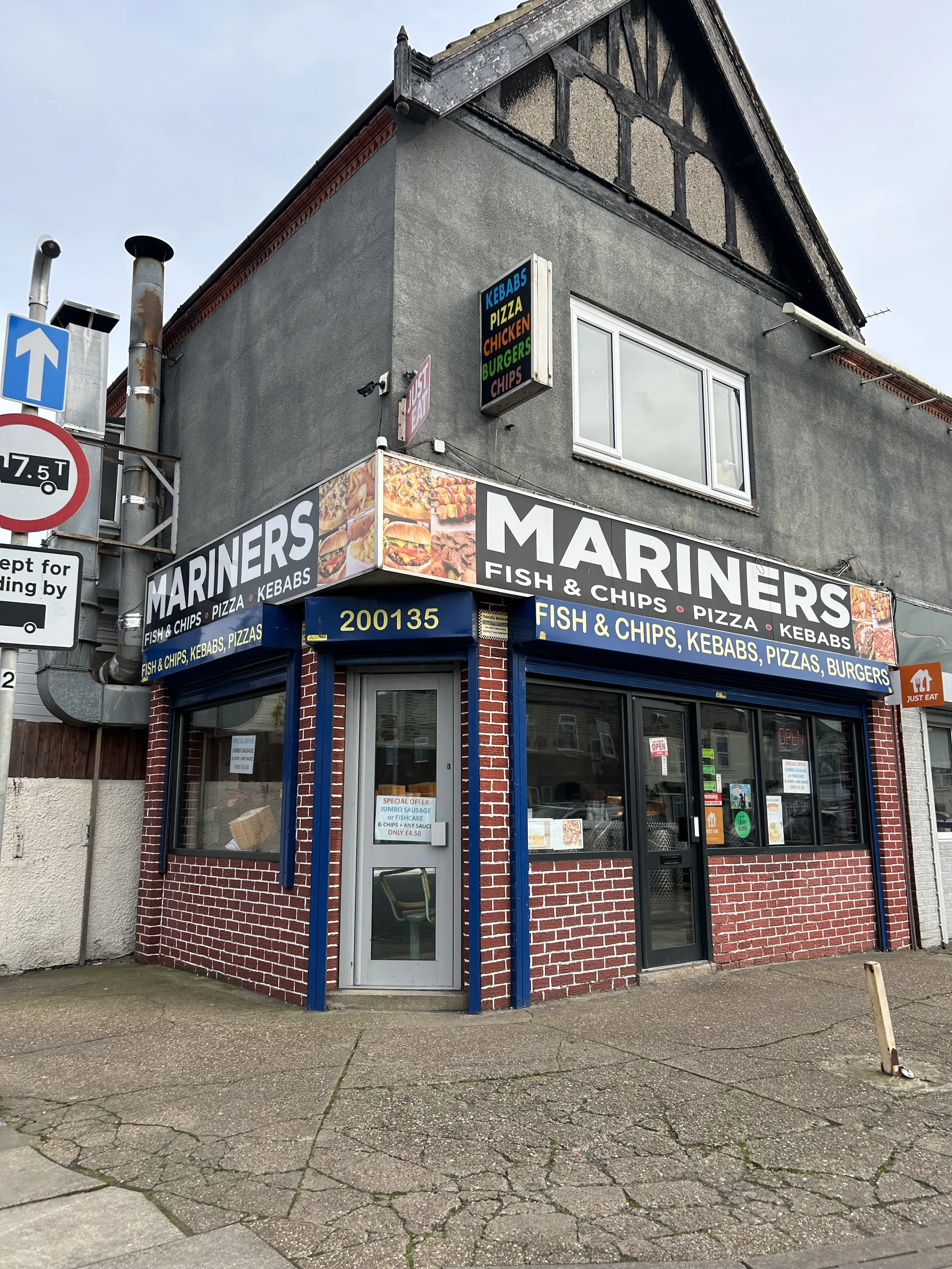 Corner view of a small restaurant named 'Mariners' offering fish and chips, pizza, kebabs, and burgers. The building has a brick and grey stucco exterior with multiple signs, including a large banner on the corner and smaller signs overhead. The stre
