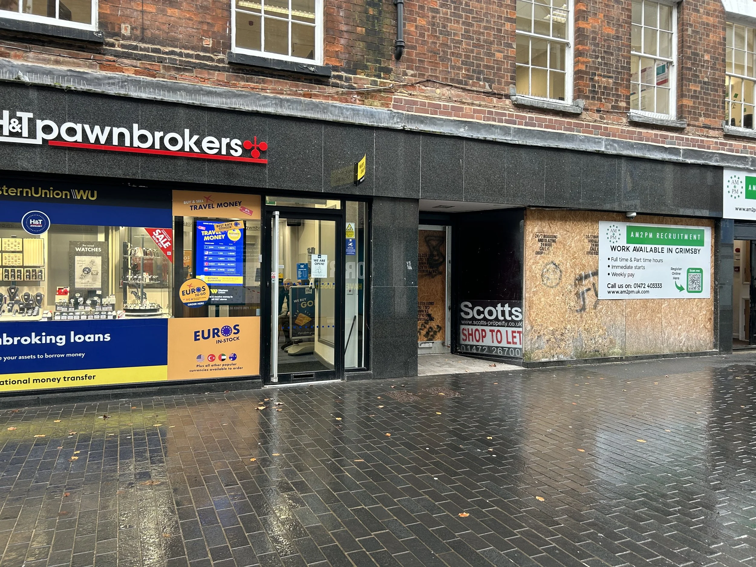 Storefront with H&T pawnbrokers sign, adjacent boarded-up shop, and a large advertisement board for AM2PM recruitment on a cloudy, rainy day in Grimsby.