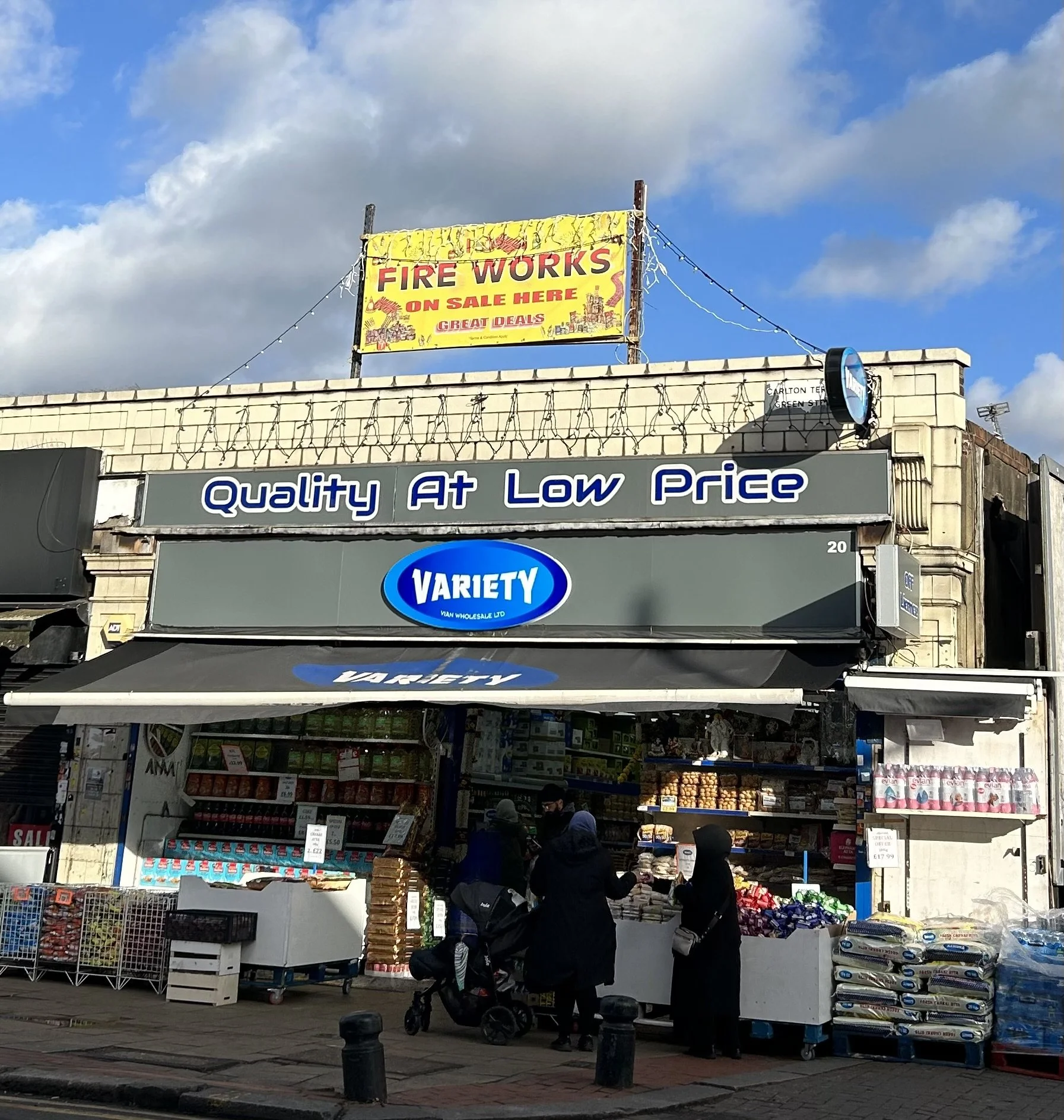 A grocery store called Variety with a sign that says 'Quality At Low Price'. There are people outside shopping, some wearing hijabs, and a yellow sign on the building advertises firework sales.