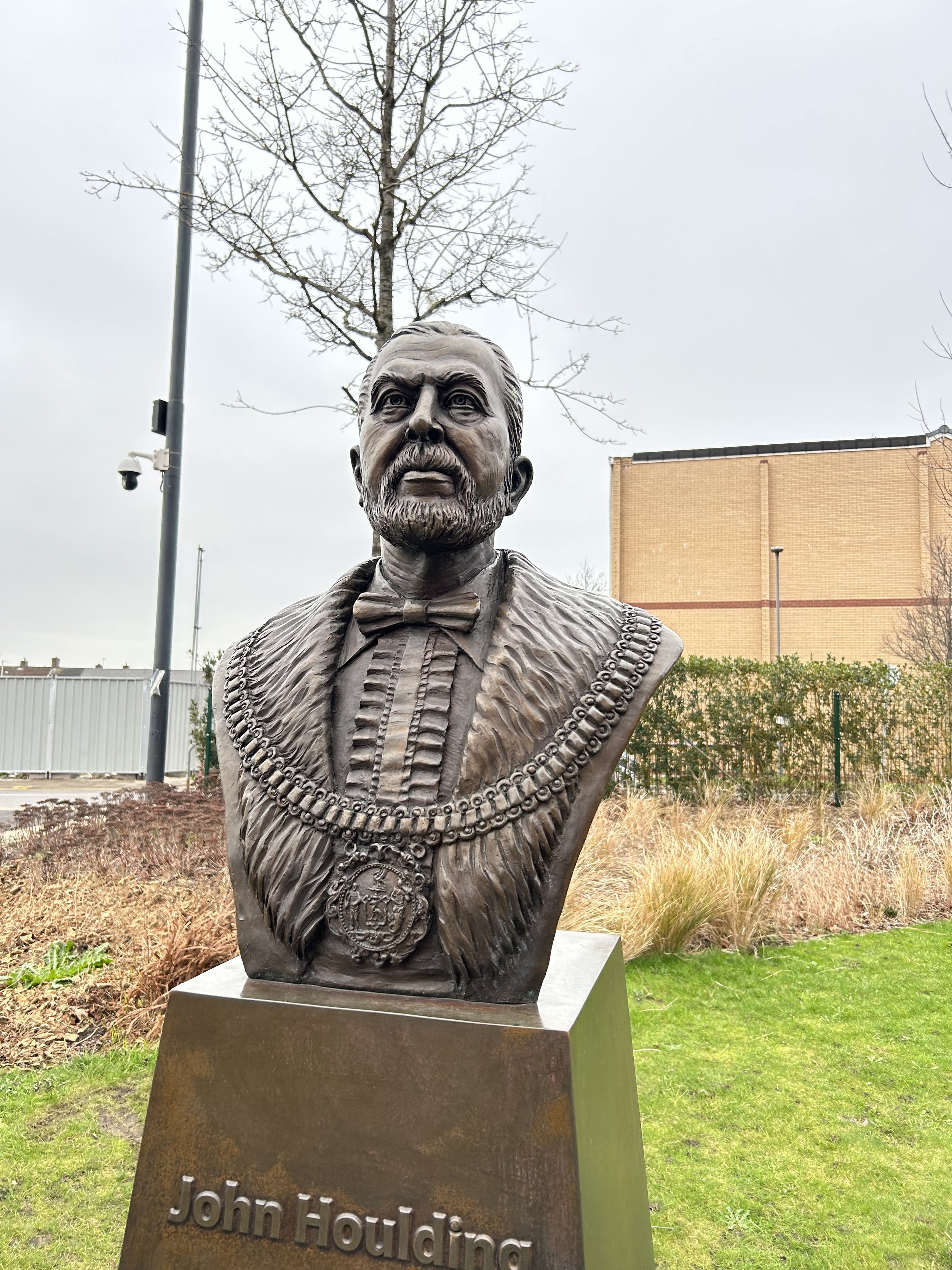 Bronze bust of John Houlding, the founder of Liverpool Football Club, with a detailed coat and medallion, outdoors on a pedestal with his name inscribed.